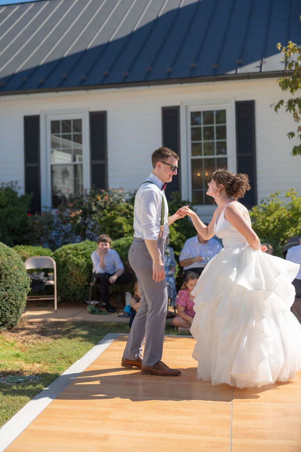 A couple shares their first dance on an outdoor wooden dance floor during a sunny backyard wedding reception.