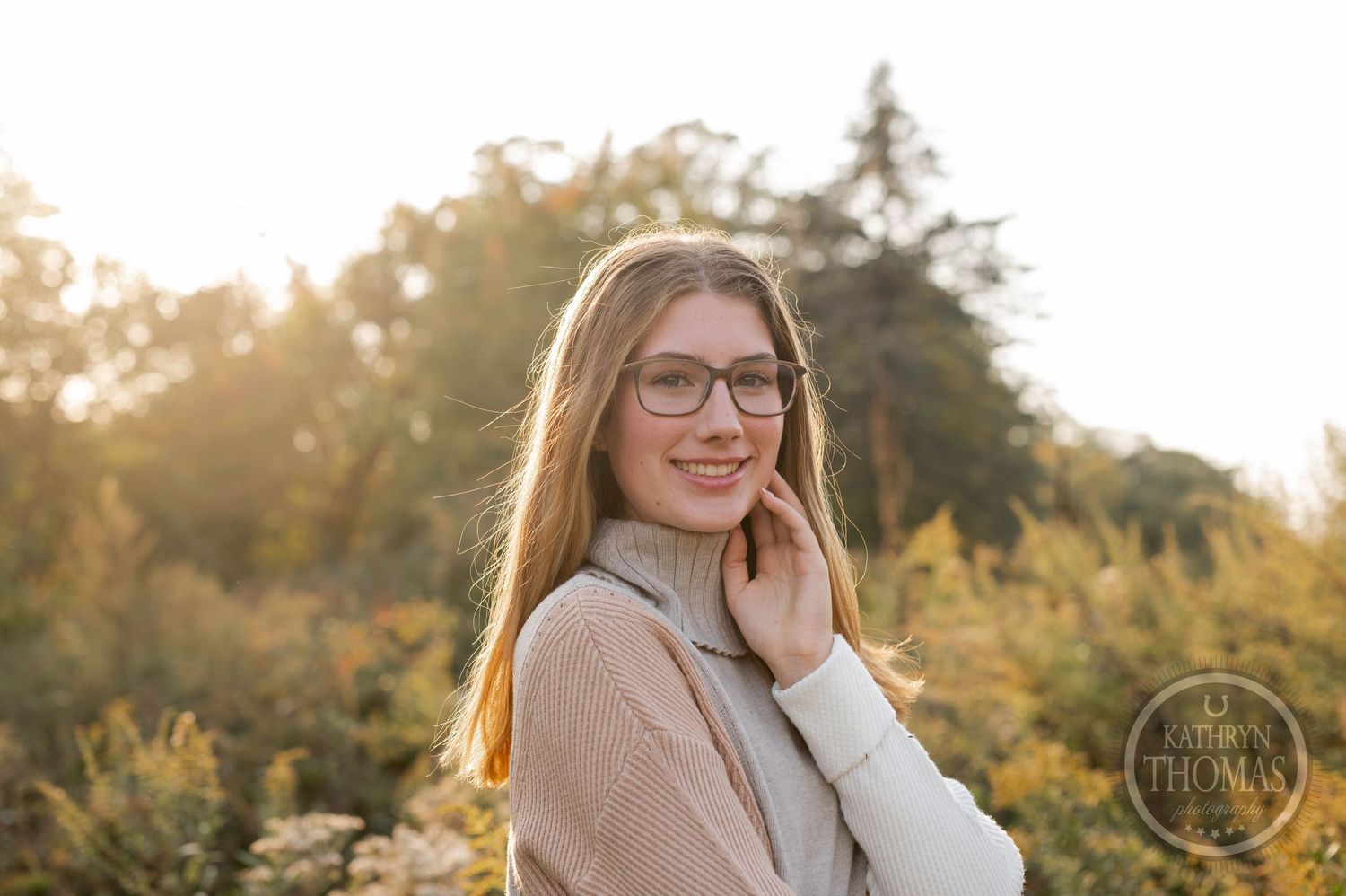 Portrait of someone in glasses and a beige sweater posing outdoors during golden hour with autumn foliage.