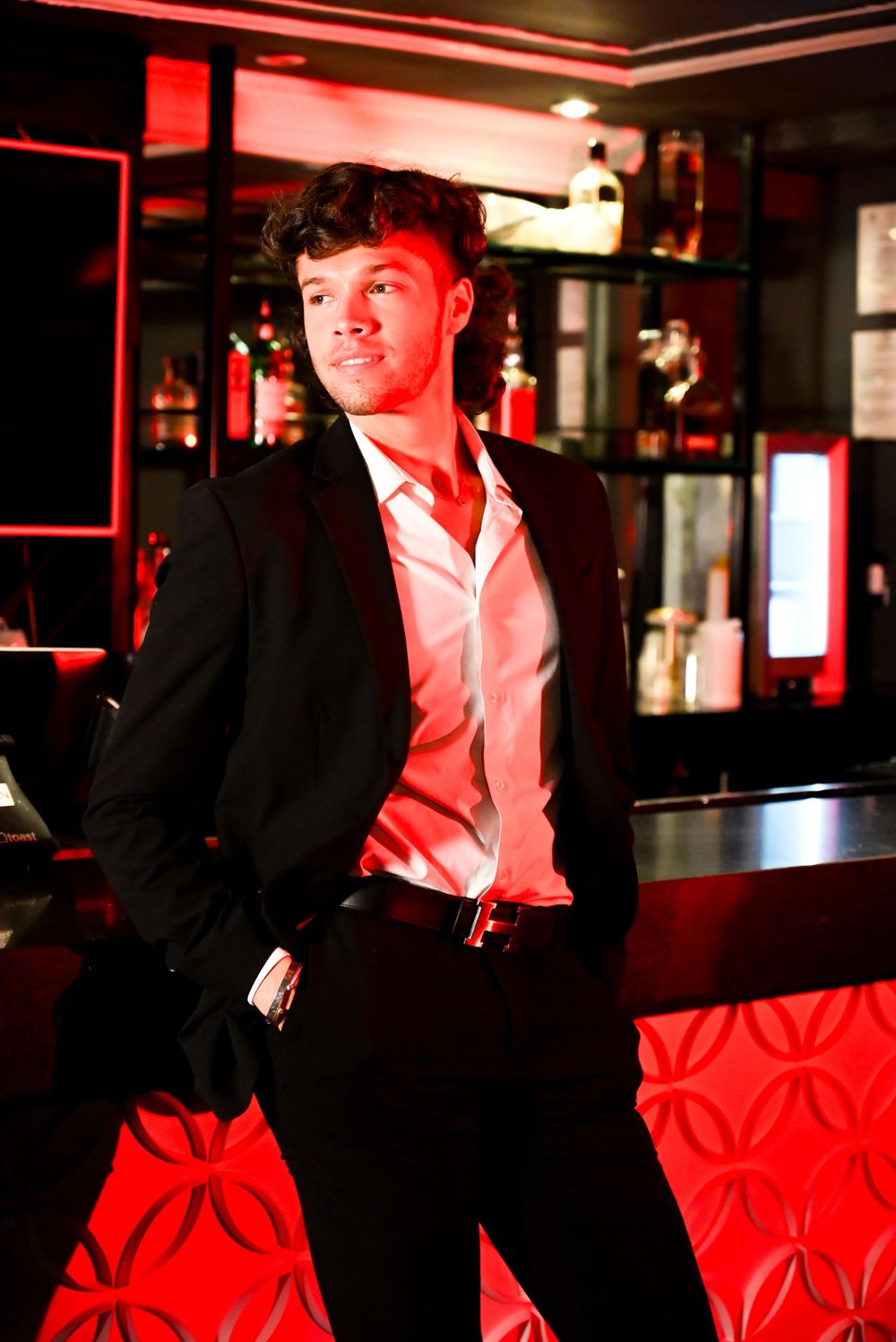 A person in a black suit and pink shirt poses against a red-lit bar interior with bottles on display in the background.