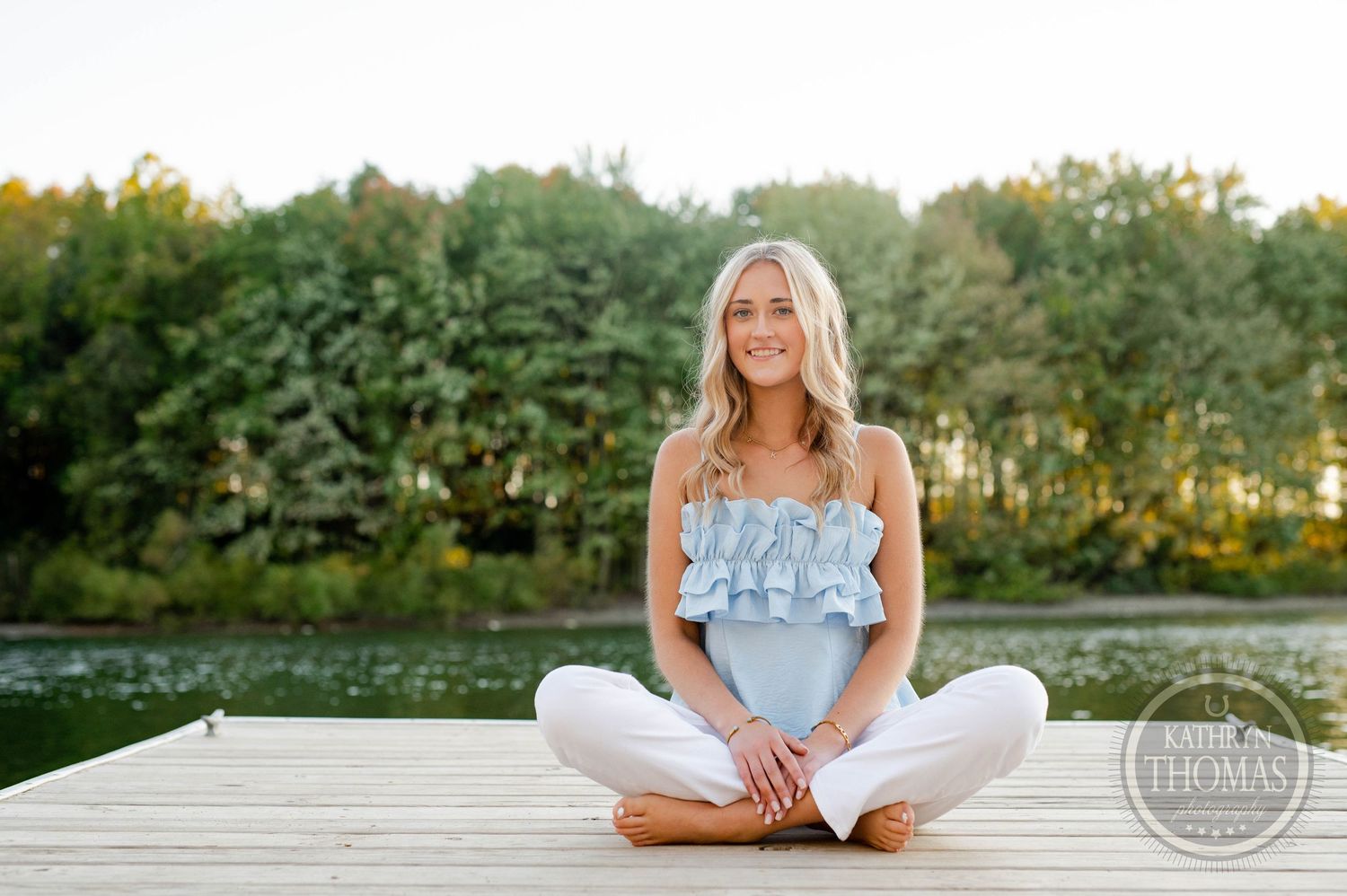 Someone in casual summer attire sitting cross-legged on a dock by a serene lake.