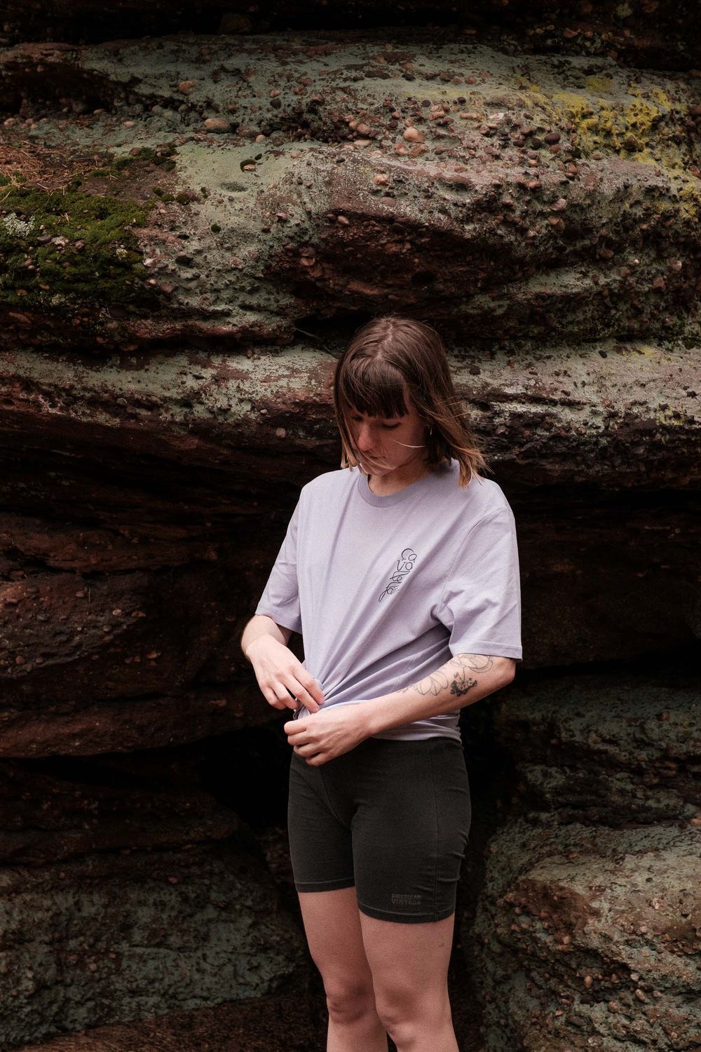 Person in light purple t-shirt standing against textured rock wall outdoors.