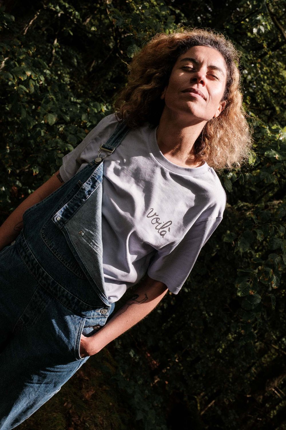 Casual outdoor portrait of someone wearing denim overalls and a gray t-shirt in natural lighting.