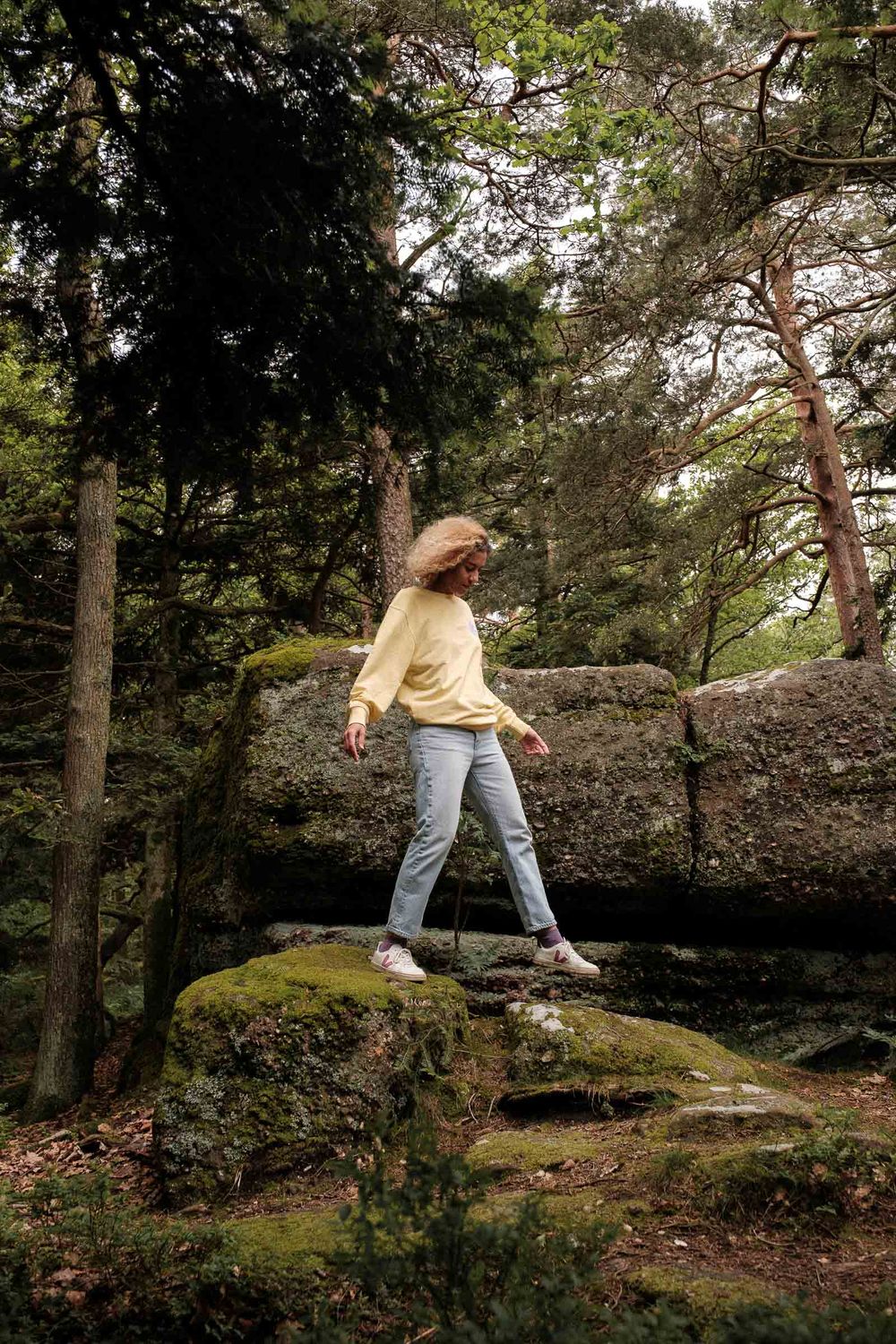 Person in yellow sweater and jeans standing on mossy rocks in forest setting.