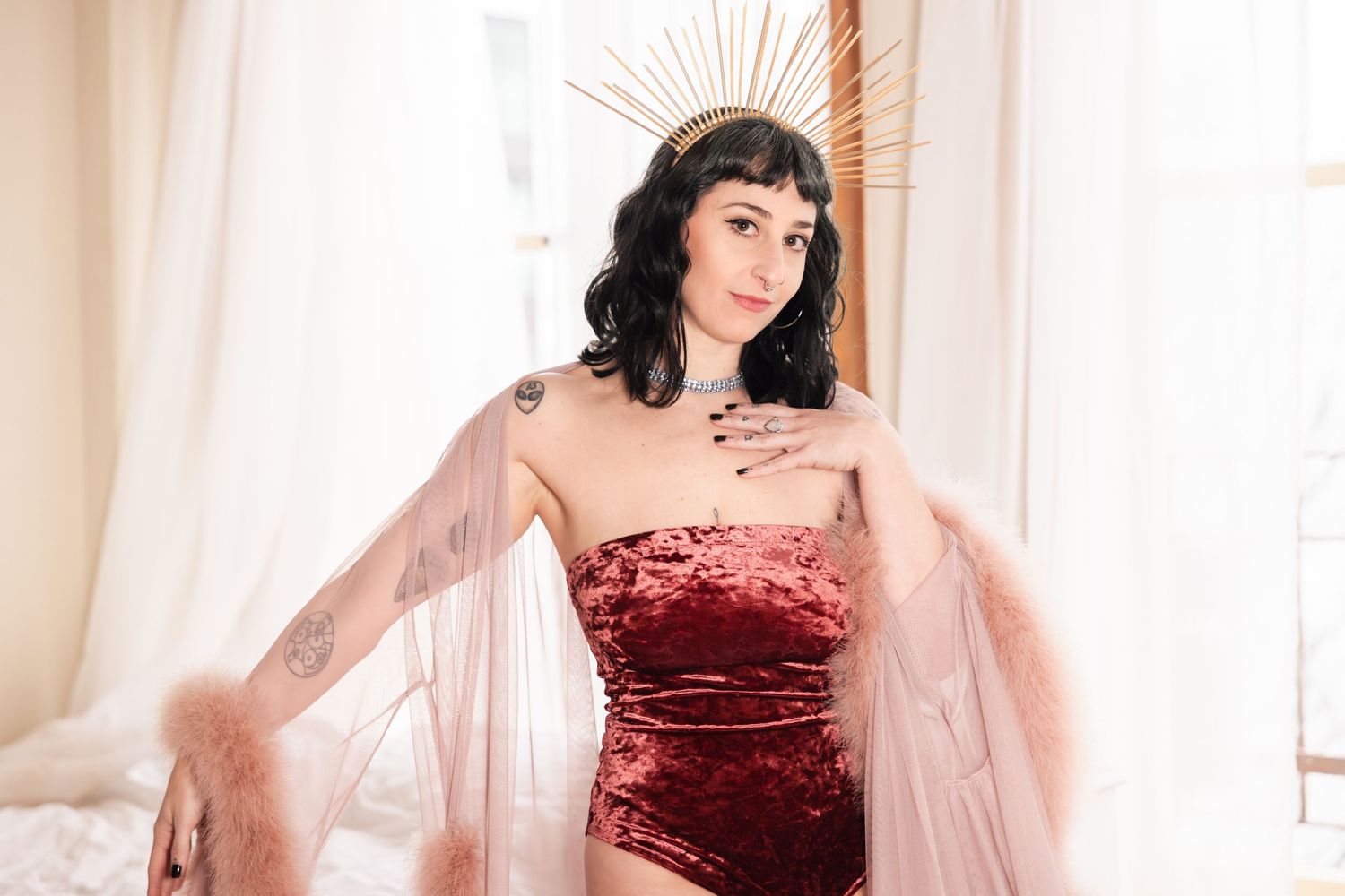 A person in a strapless red velvet dress and sheer pink cape poses near light window curtains.