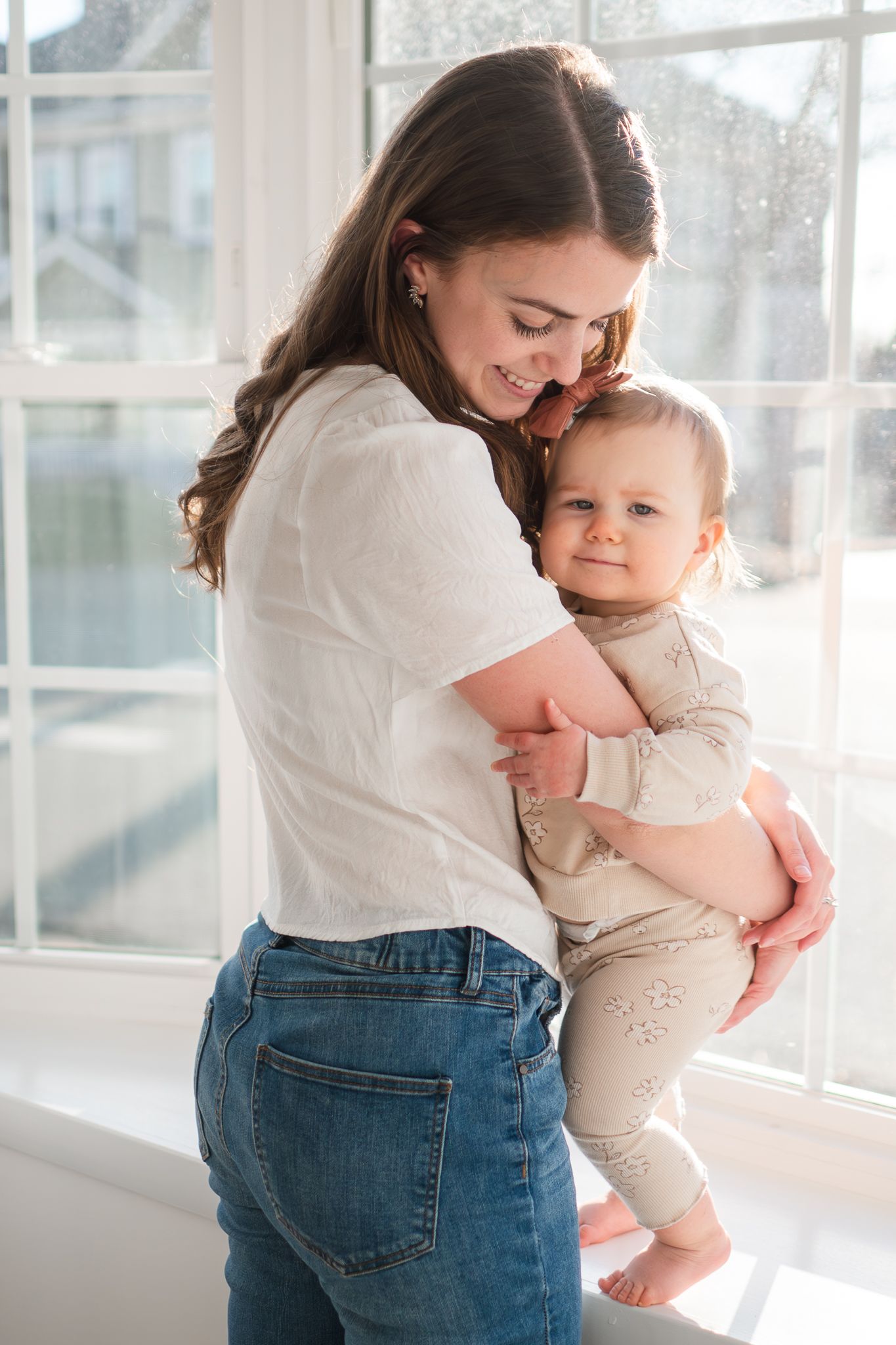A tender moment between a woman in jeans and white top embracing a baby wearing beige by a bright window.