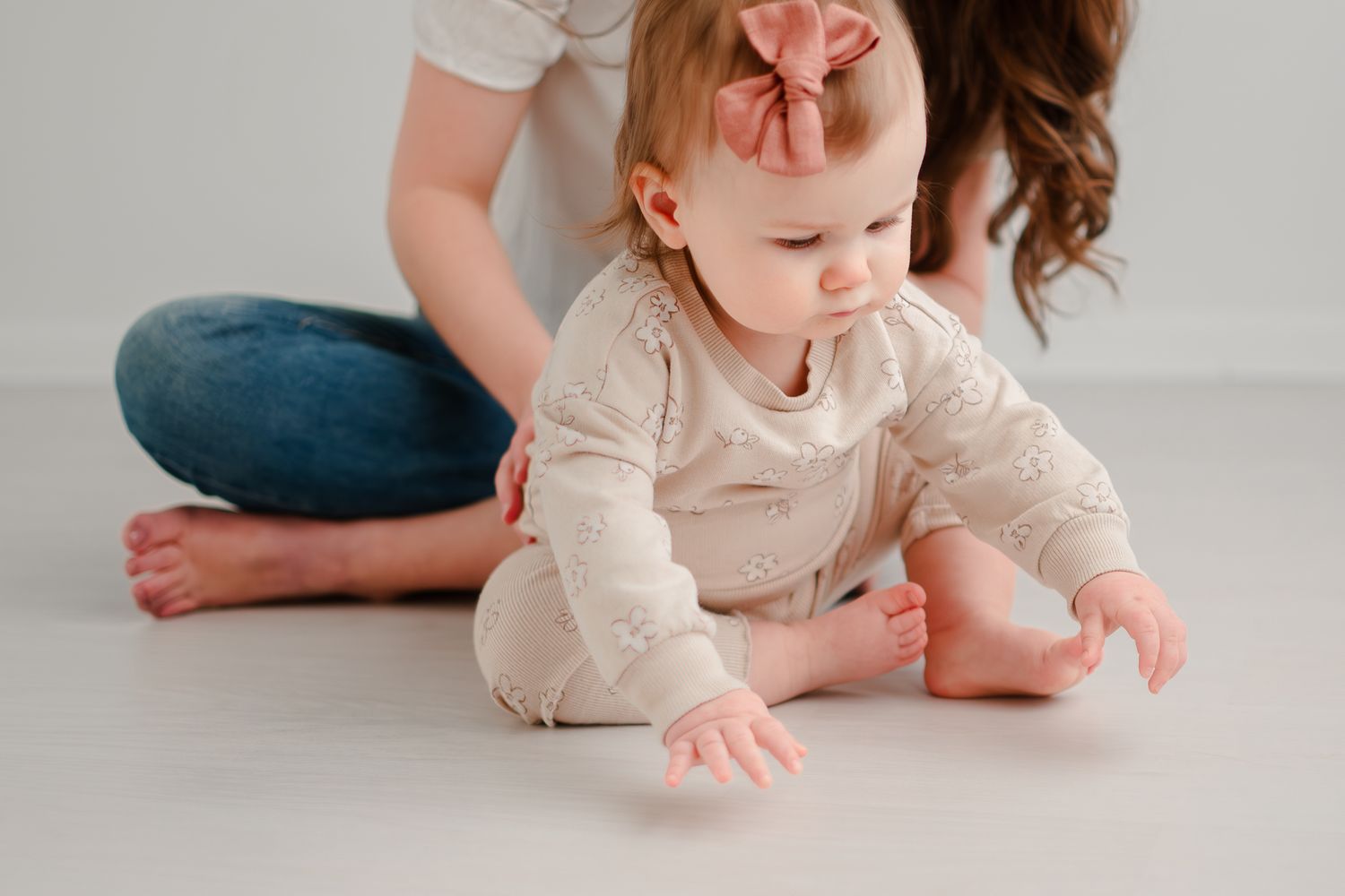 A baby wearing a cream-colored sweater and peach bow crawls on a light floor while someone sits nearby.
