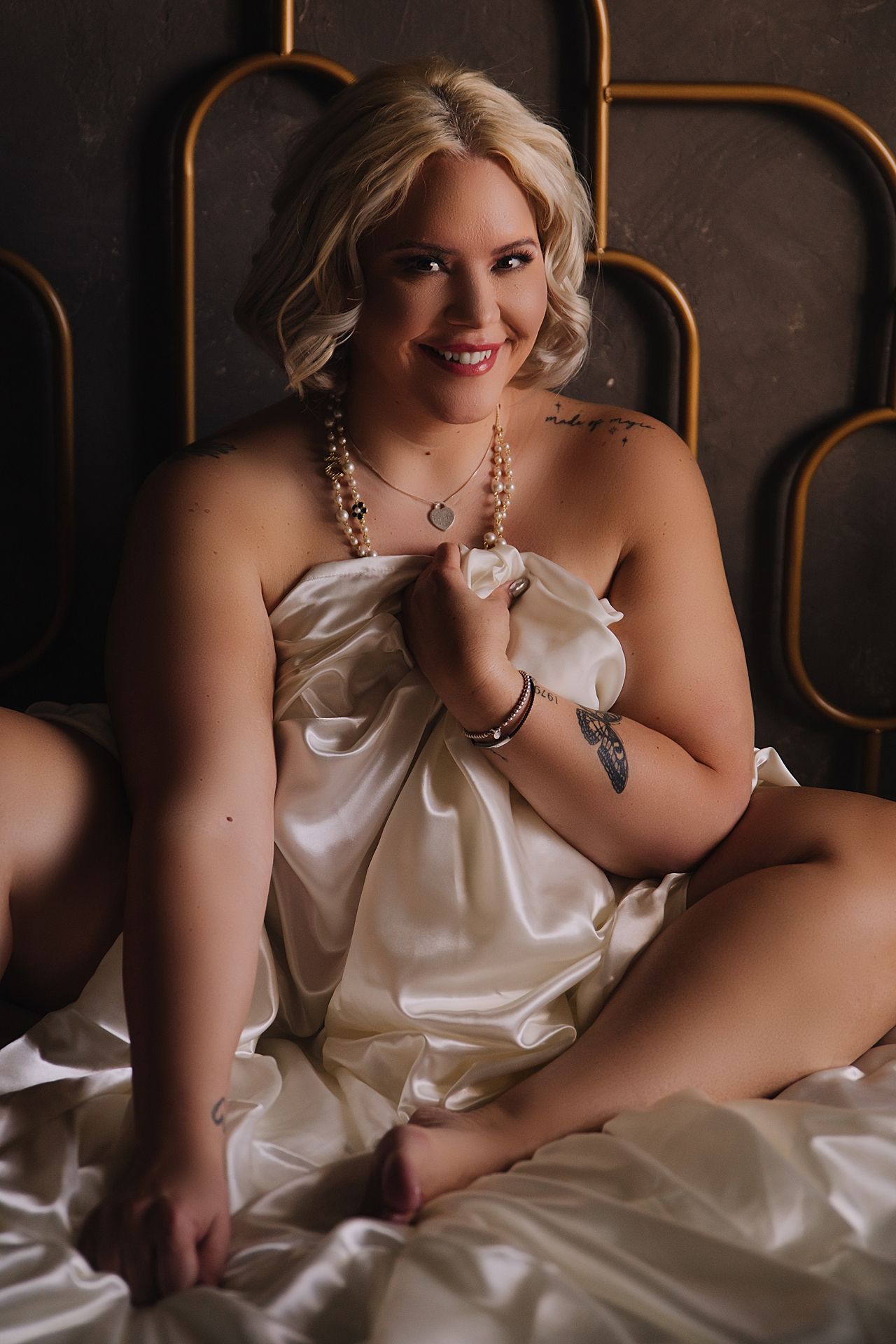 Artistic boudoir portrait featuring white satin fabric and pearl necklace in moody lighting with warm brown accents.