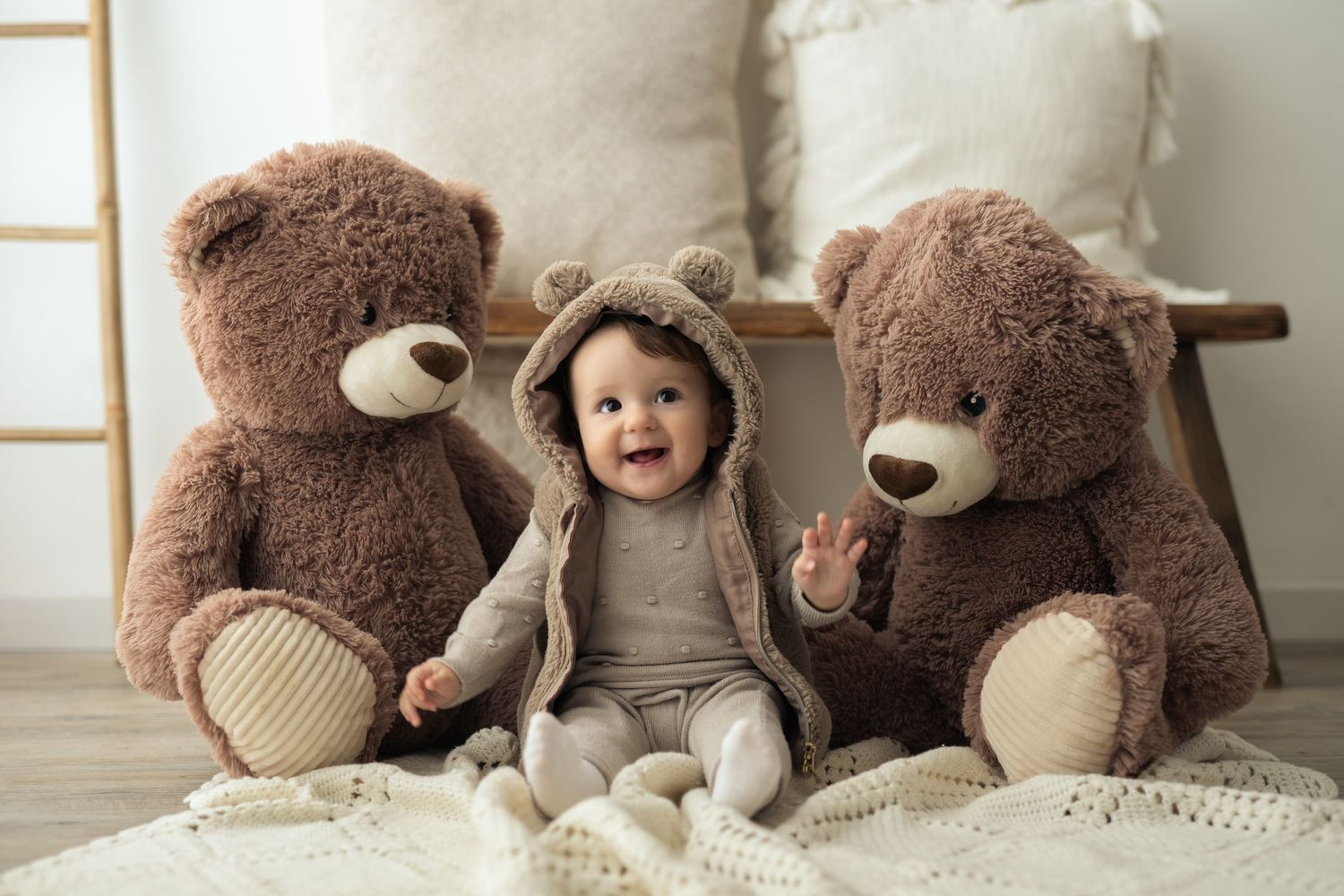 Bambina di un anno con cappuccio di pelo e orecchie da orso, sorride seduta su una coperta beige e sullo sfondo due orsi di peluches