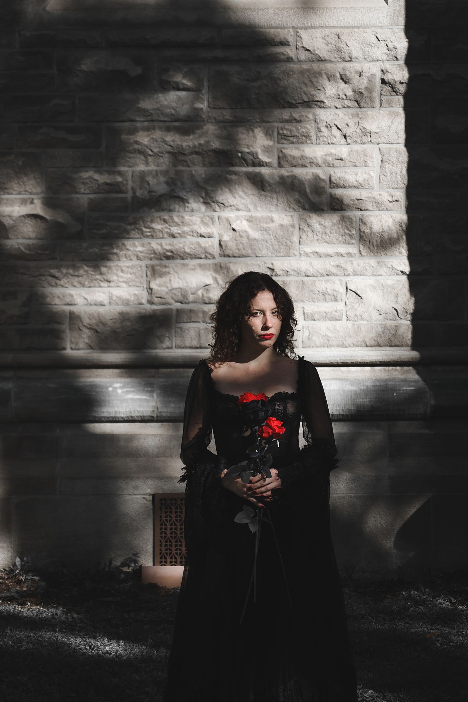 Woman in a black flowing dress holds red roses against a stone wall with dramatic shadow play and moody lighting.