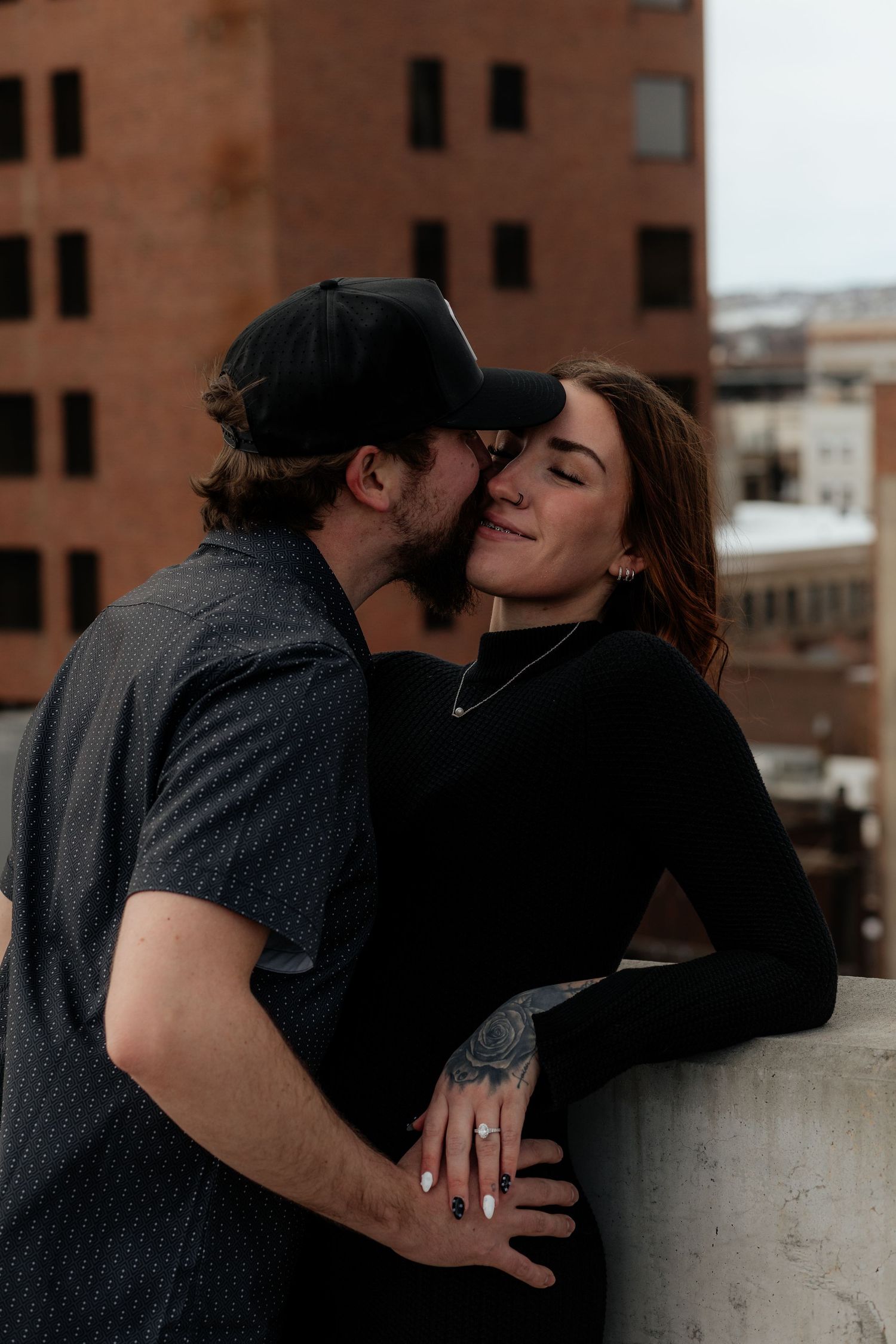 A couple shares an intimate moment on a rooftop with brick buildings in the background at dusk.