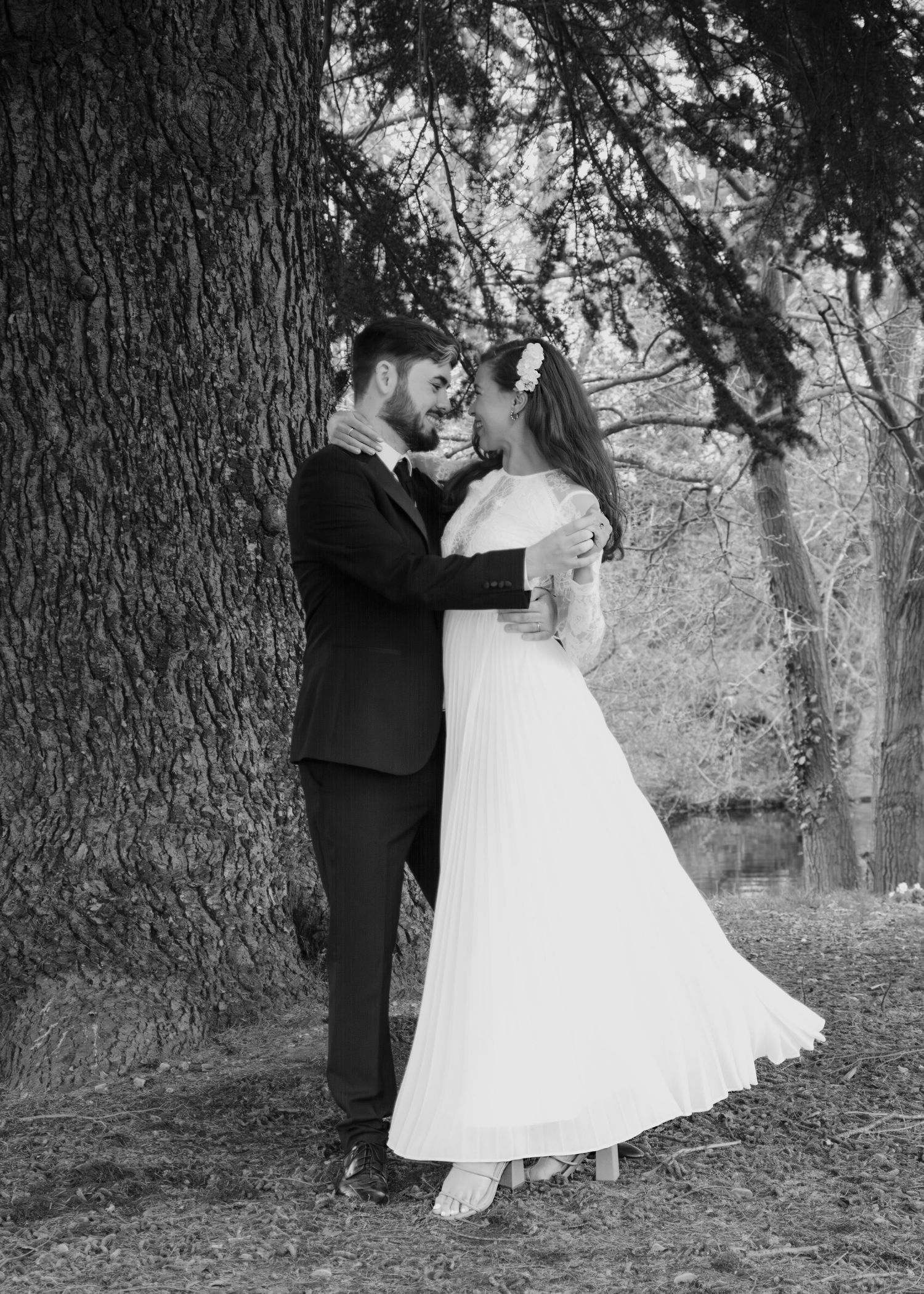 A bride and groom share a romantic embrace beside a large tree in a black and white wedding portrait.