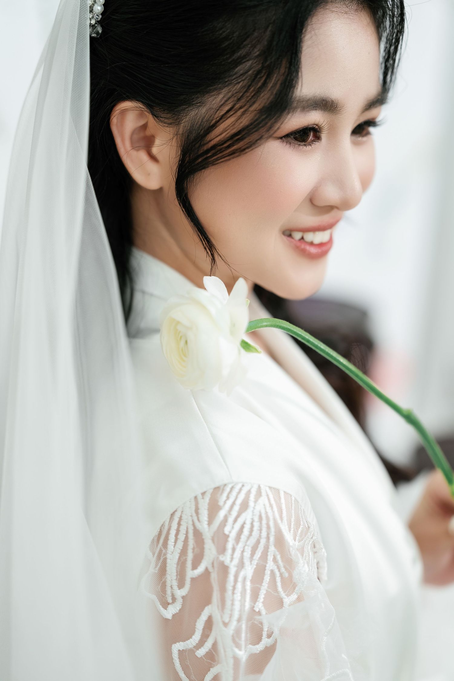Side profile view of a bride in white lace wedding dress holding a cream colored flower against a light background.