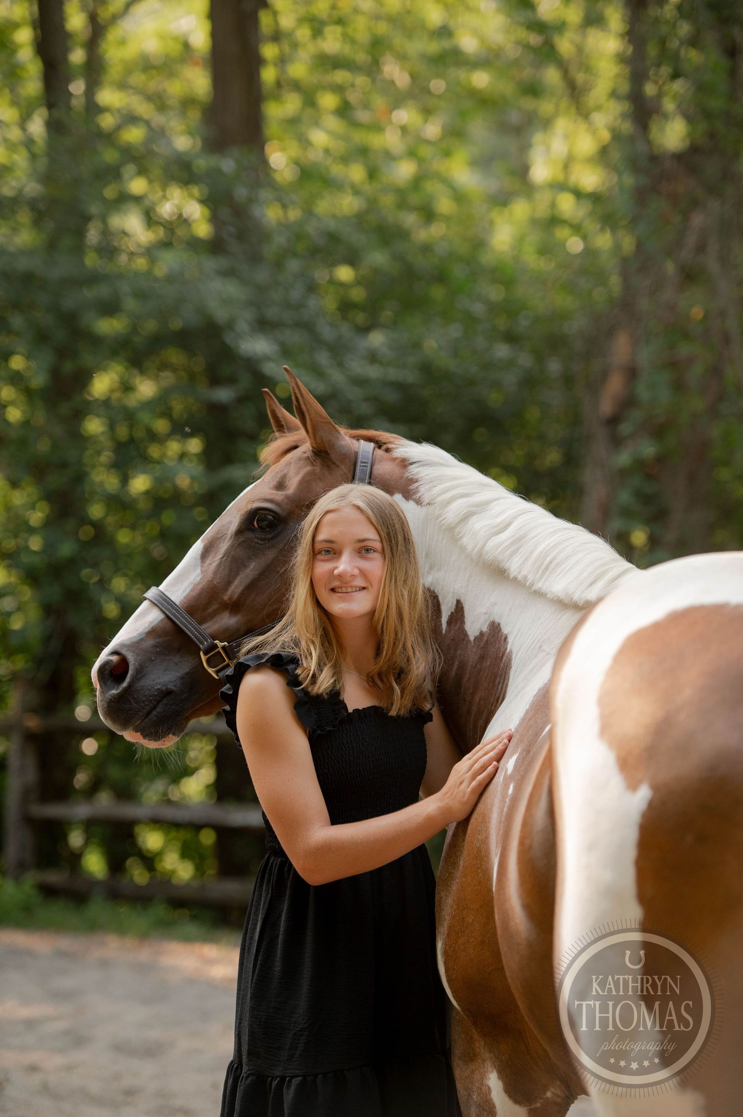 A person wearing a black dress hugs a brown and white horse while standing in a forest clearing.