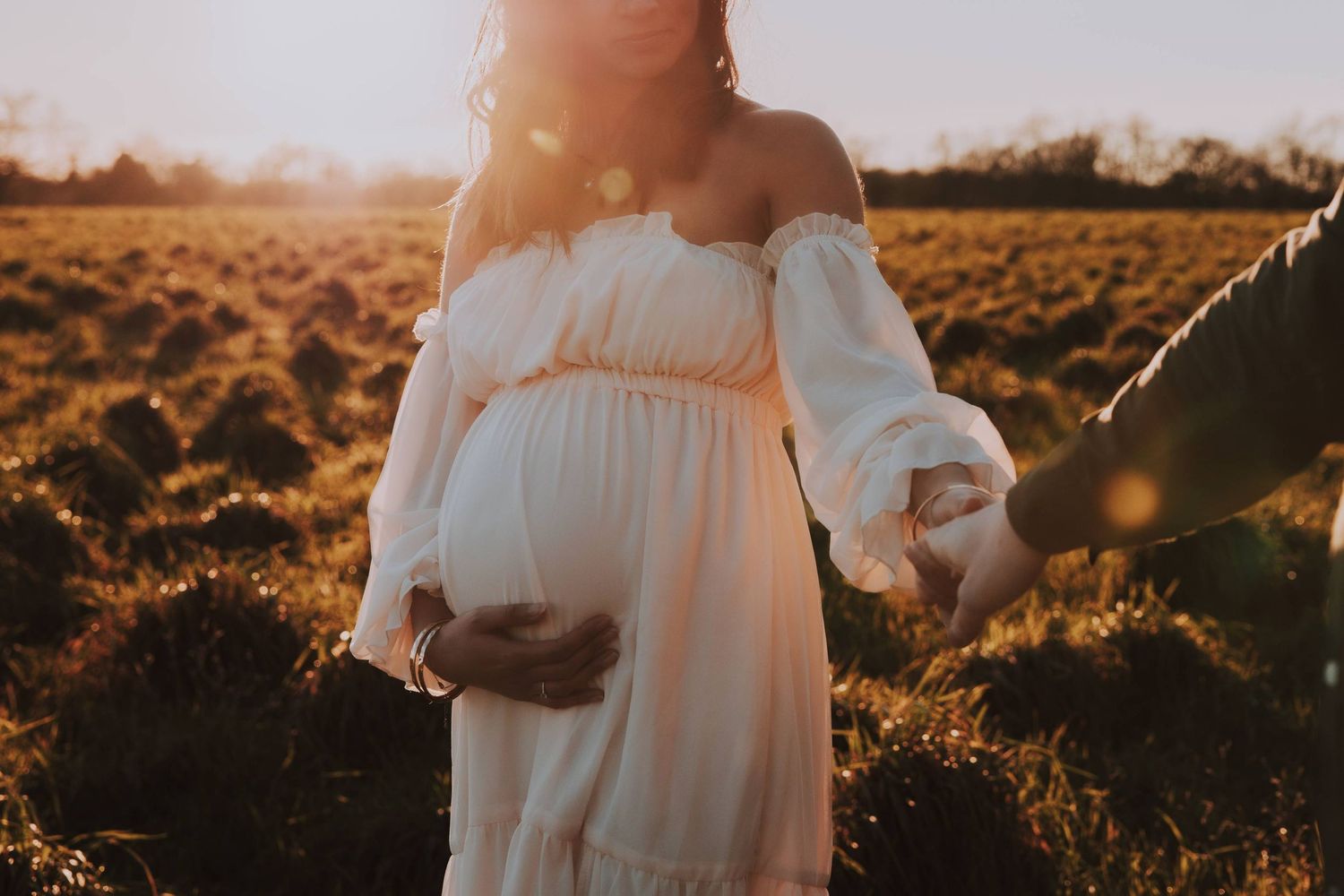 A pregnant woman in a flowing white dress stands in a golden field at sunset.