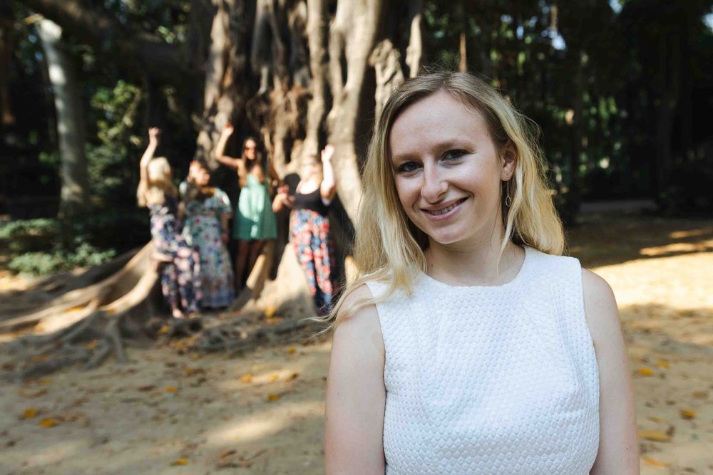 Portraits de groupe EVJF à Sévill
A person in a white sleeveless top stands outdoors with others visible in the background near large trees.
