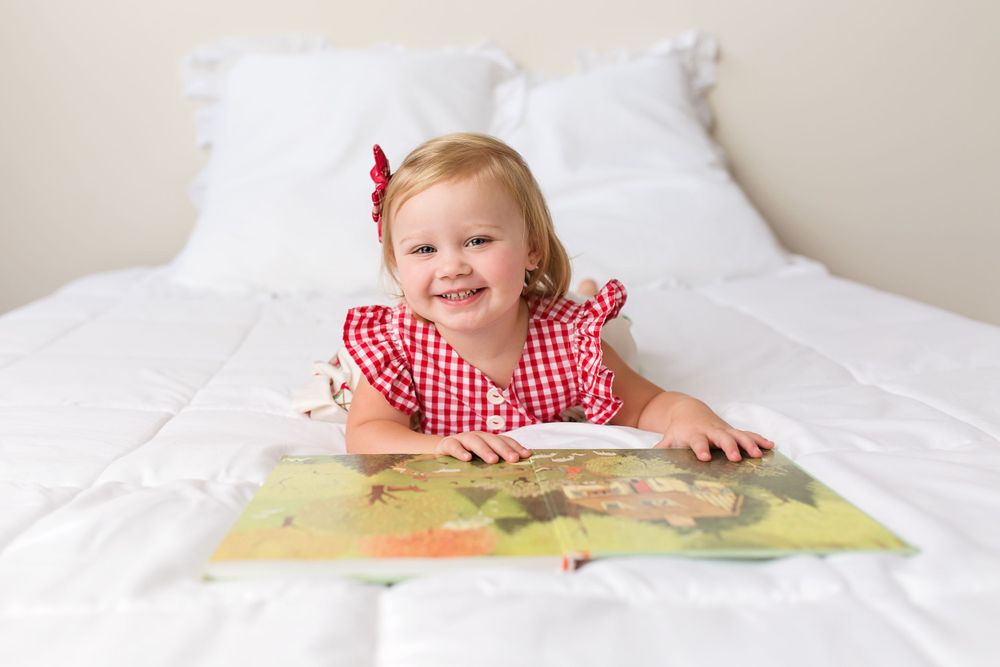 A child in a red gingham dress reading a picture book while lying on white bedding.