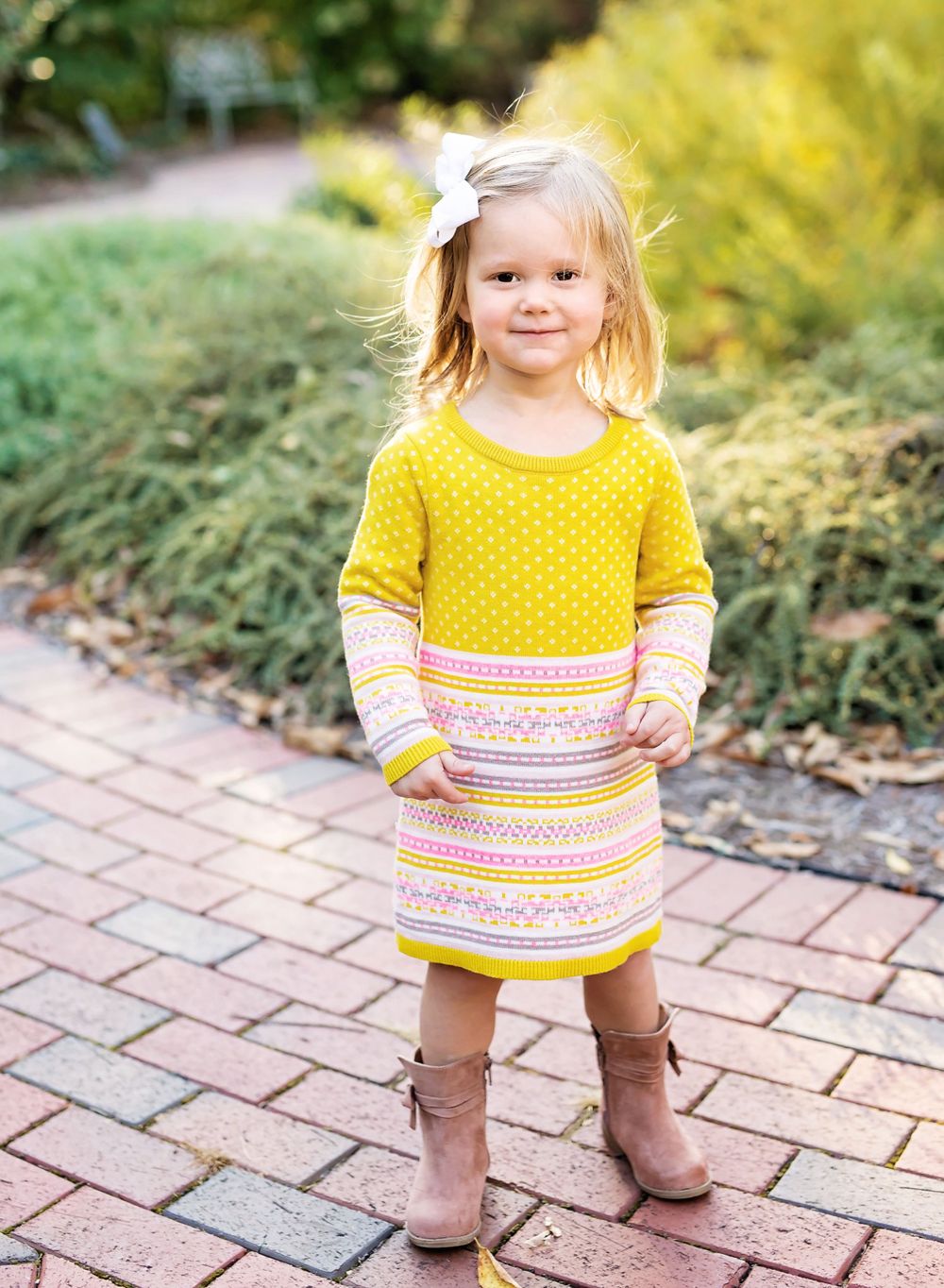 Child wearing yellow patterned dress and brown boots standing on brick pathway in garden setting.