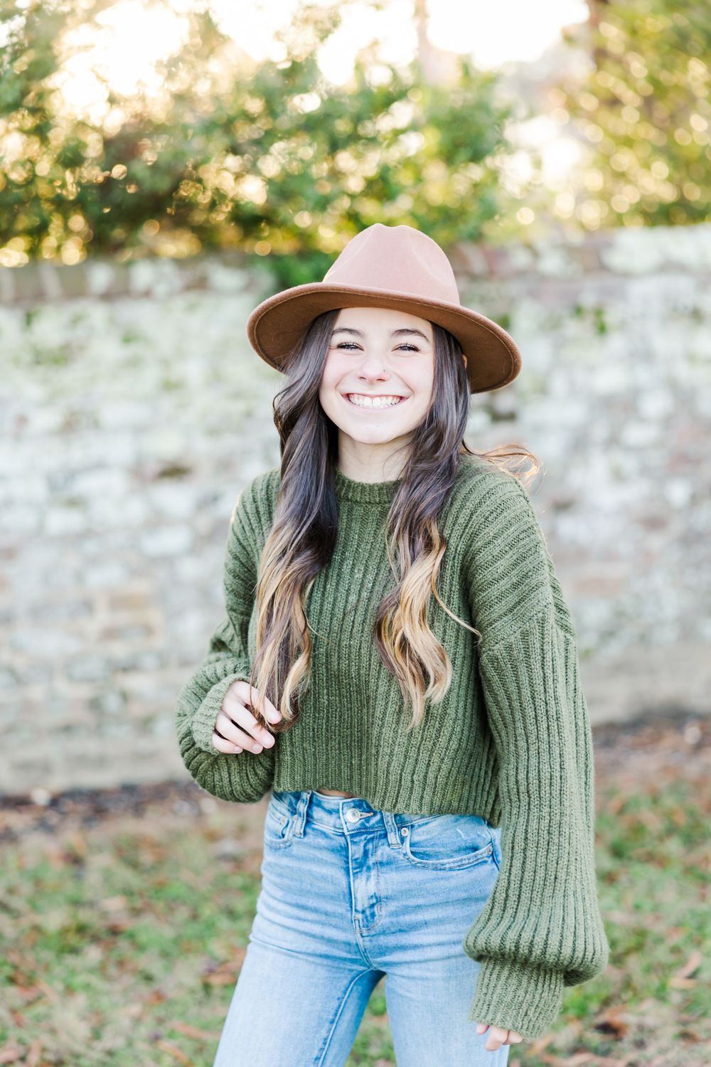 Person wearing a green sweater, brown hat and jeans poses outdoors on a sunny day.