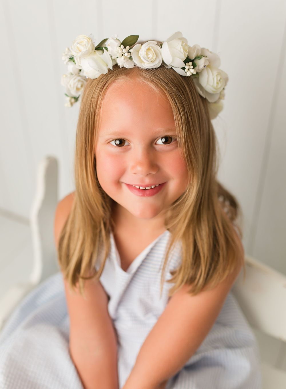 Child wearing white floral crown and dress poses against light background.