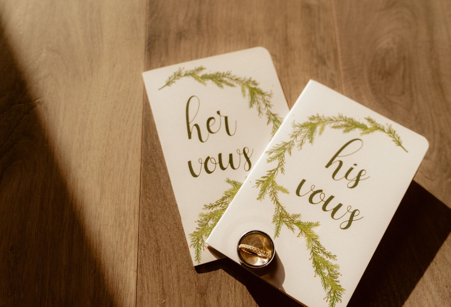 Wedding rings rest between two white cards with elegant calligraphy reading 'her vows' and 'his vows' on wooden surface.
