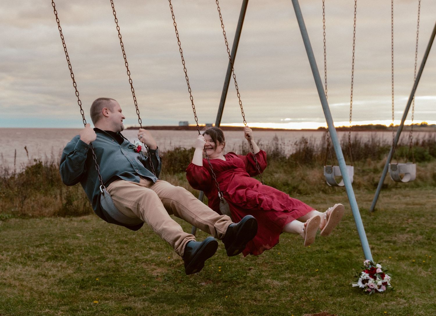 Two people enjoying swings at sunset with the ocean in the background and tall grass in the foreground.