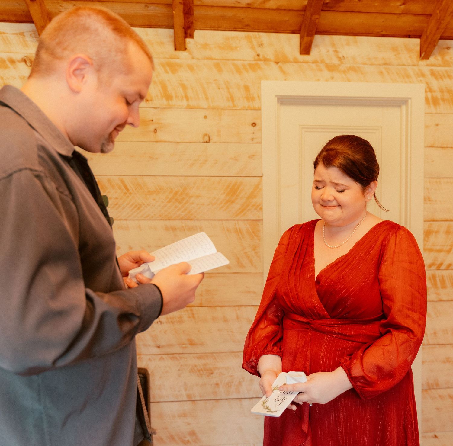 Someone in a red wrap dress and another person in gray both look down at papers in a wood-paneled room during a ceremony.