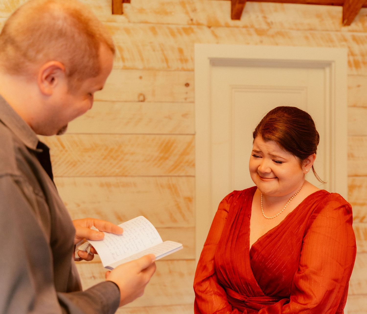 Two people sharing a moment at a rustic cabin venue, one writing in a book while wearing gray as the other in red smiles warmly.