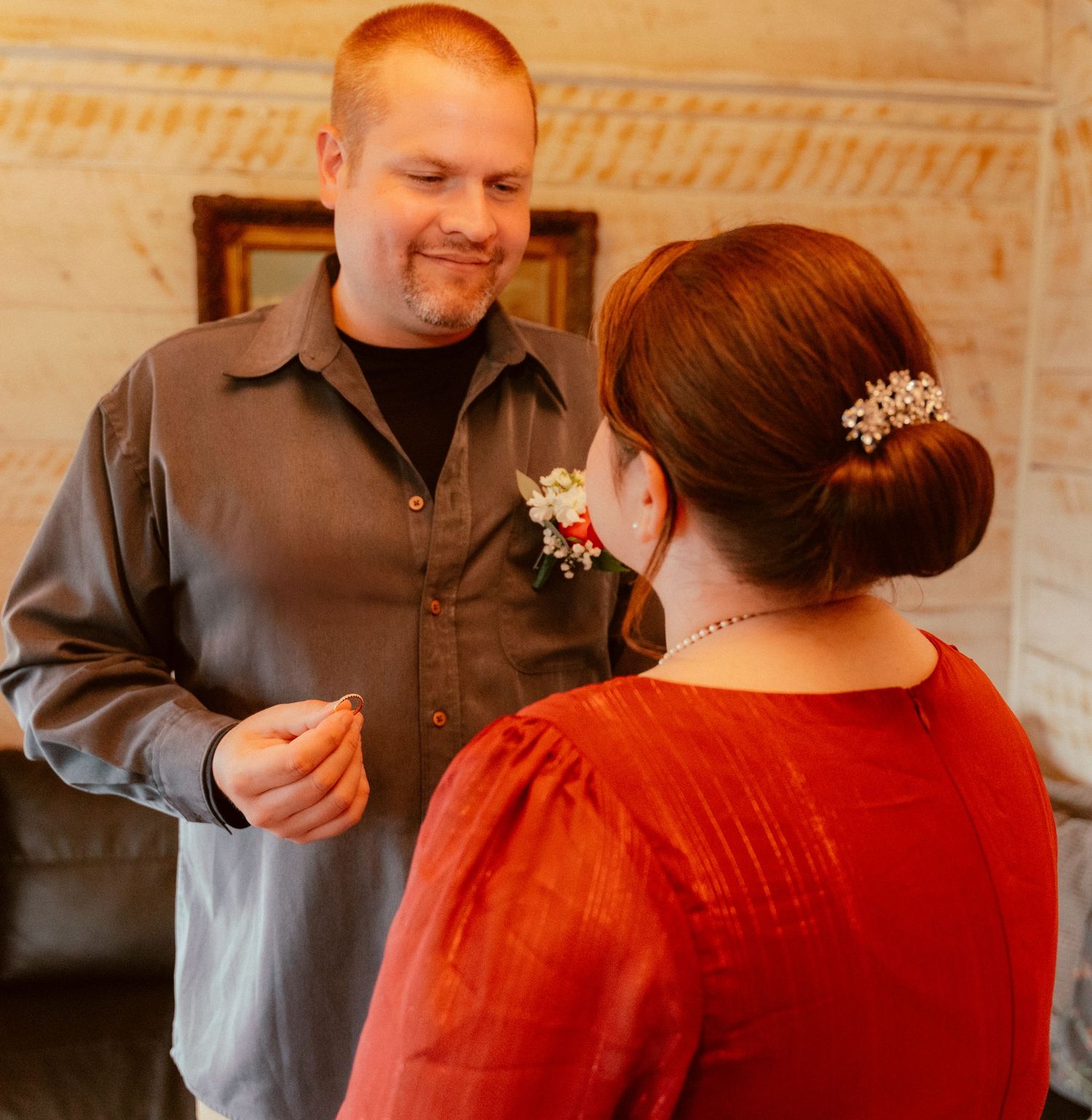 A couple shares a tender moment indoors, with one wearing a brown shirt and the other in a red dress with hair ornaments.