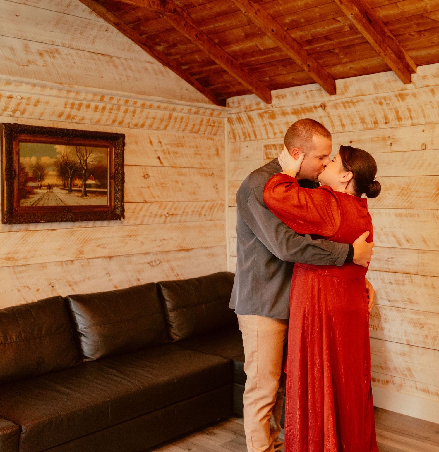 A tender embrace between two people wearing red and gray standing in front of a dark couch in a wooden interior.