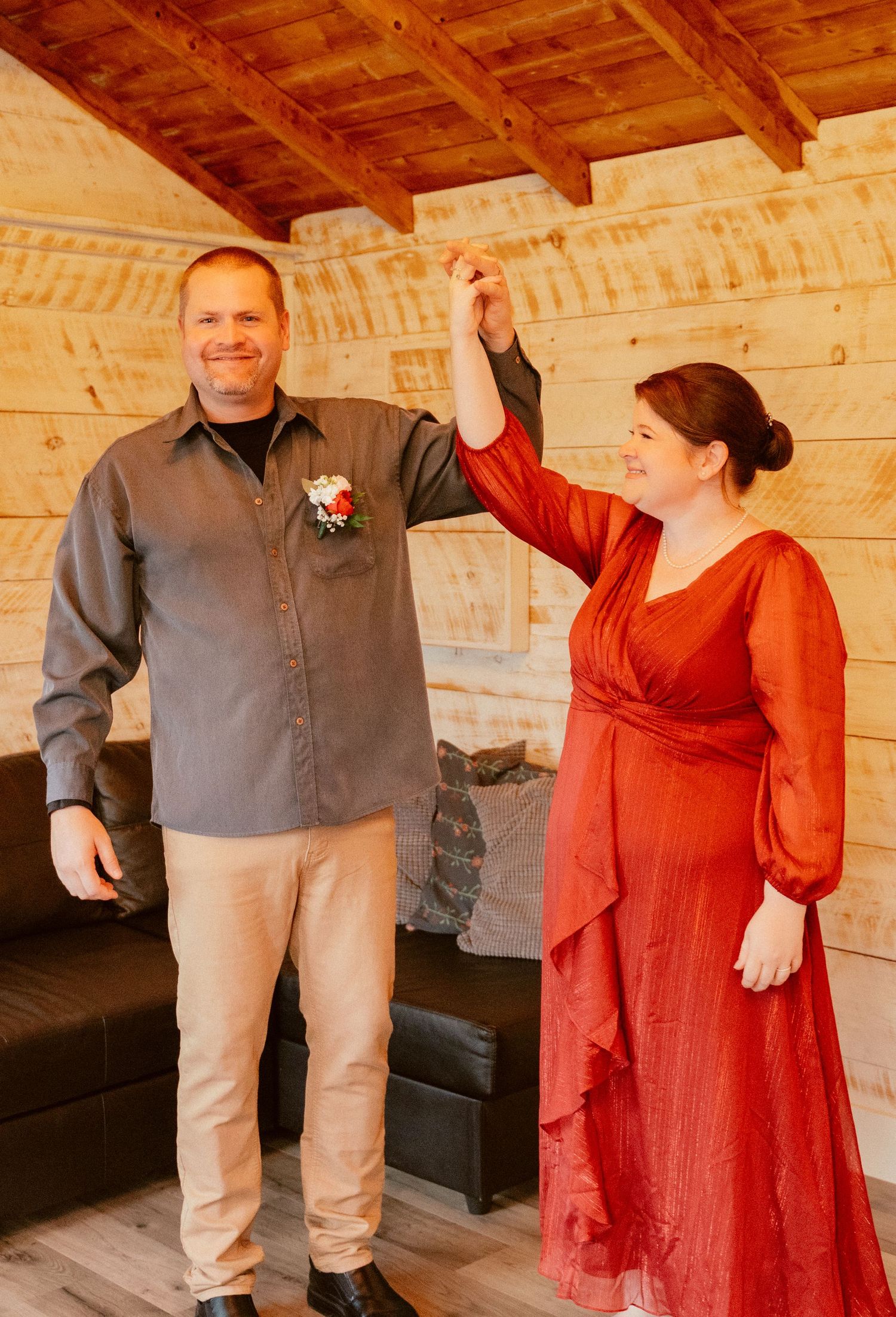A couple in a rustic wood-paneled room, wearing a gray shirt and flowing red dress during a special occasion.