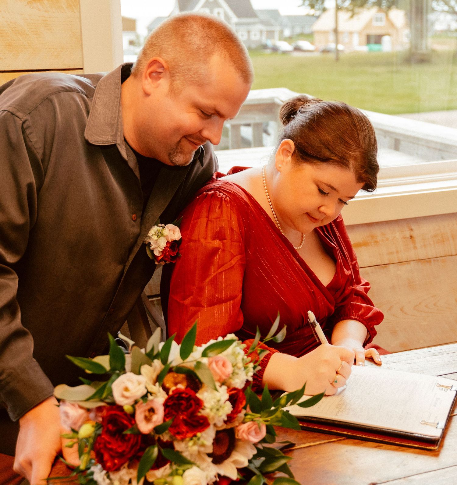 A couple signs marriage documents beside a beautiful floral bouquet near a window.