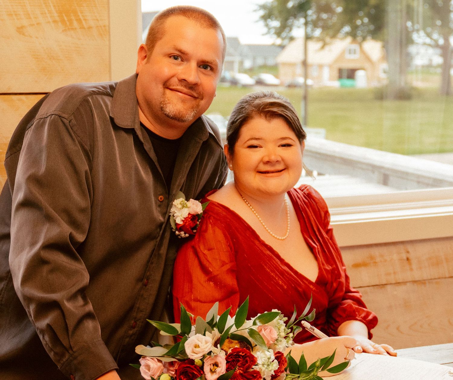 A couple in formal wear smiling together with floral decorations at an indoor celebration.