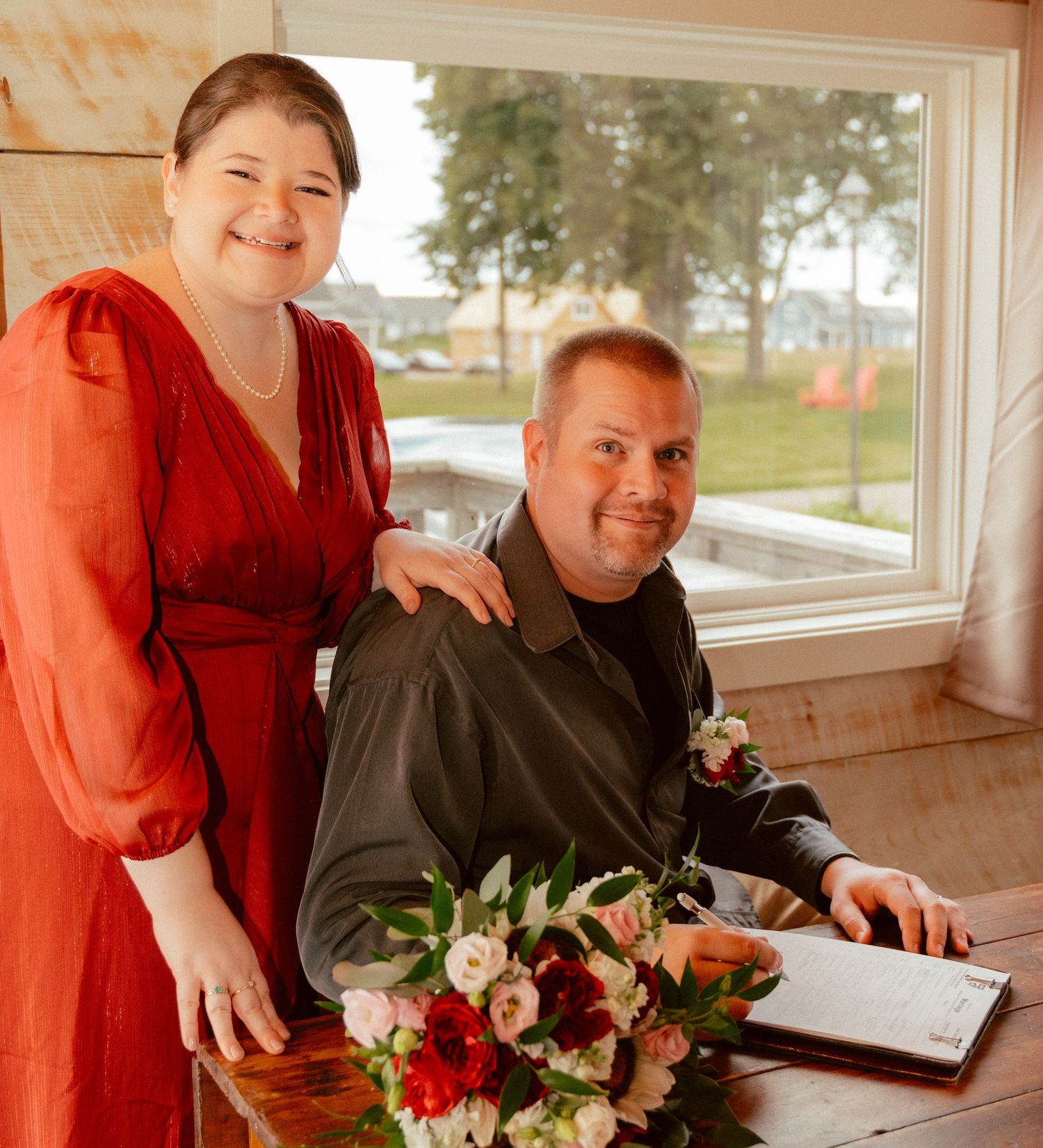 A happy couple poses together at their wedding with a vibrant bouquet on a wooden table.