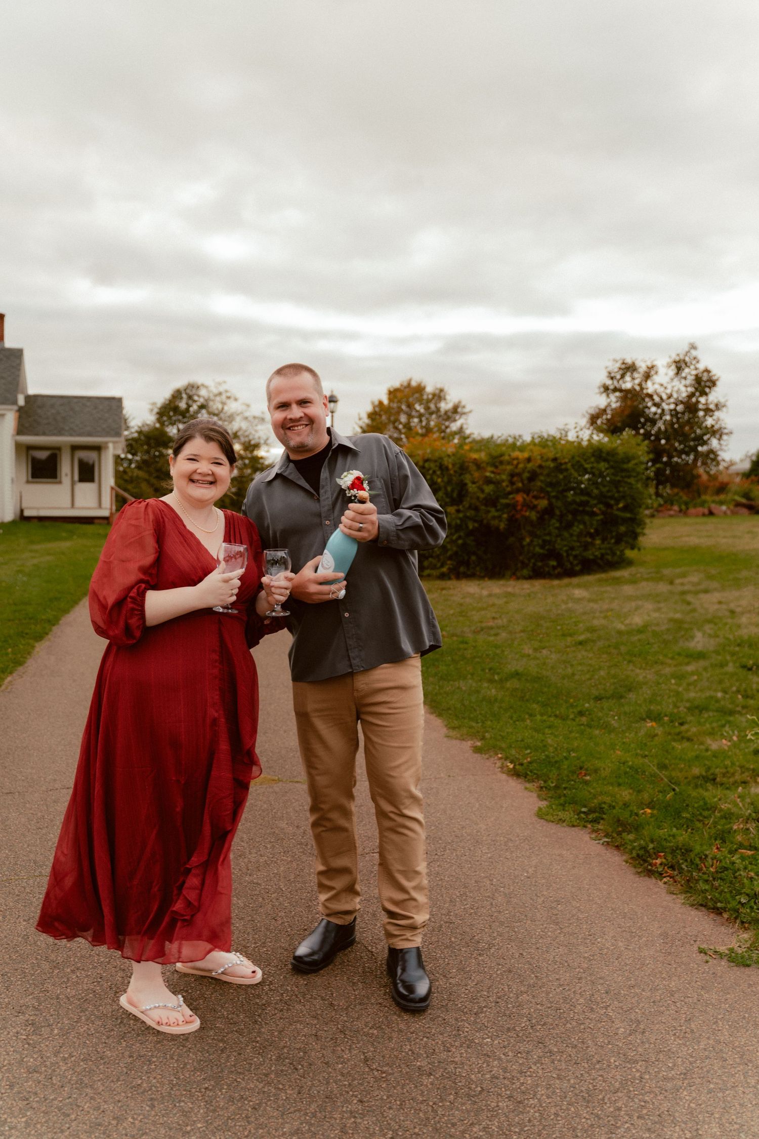 Couple in formal attire holding drinks while standing on pathway with house in background.