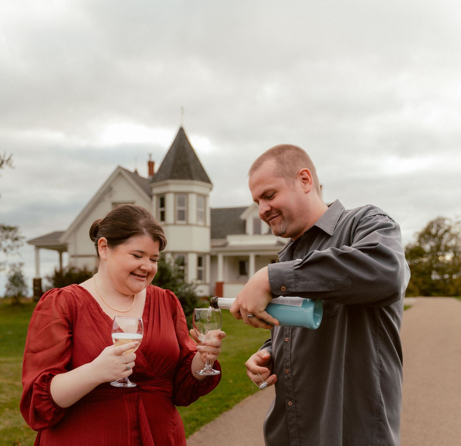 A couple shares a joyful moment pouring drinks outside a Victorian house on a cloudy day.