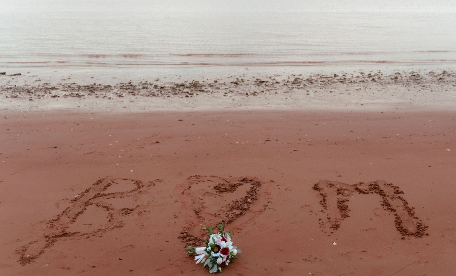 A flower bouquet lies on red beach sand next to letters written in the sand.