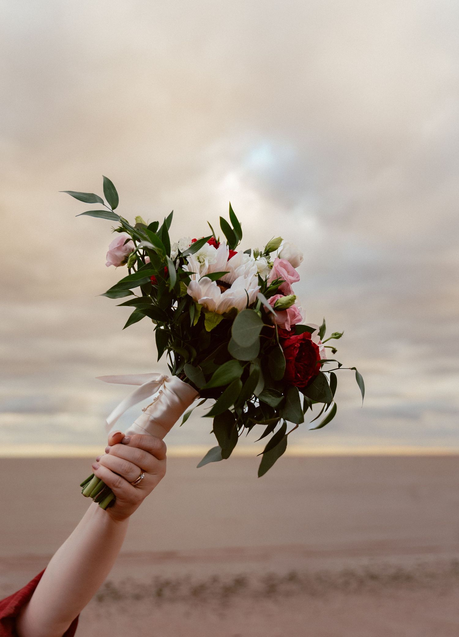A hand holds up a beautiful bouquet of white and red flowers against a cloudy sunset sky.