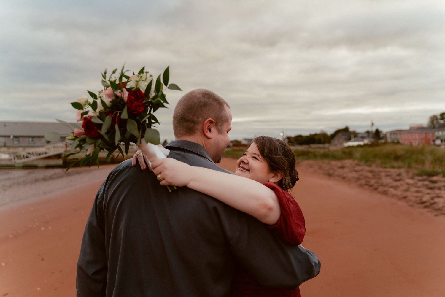 A couple embraces on a red sandy beach during an overcast evening.