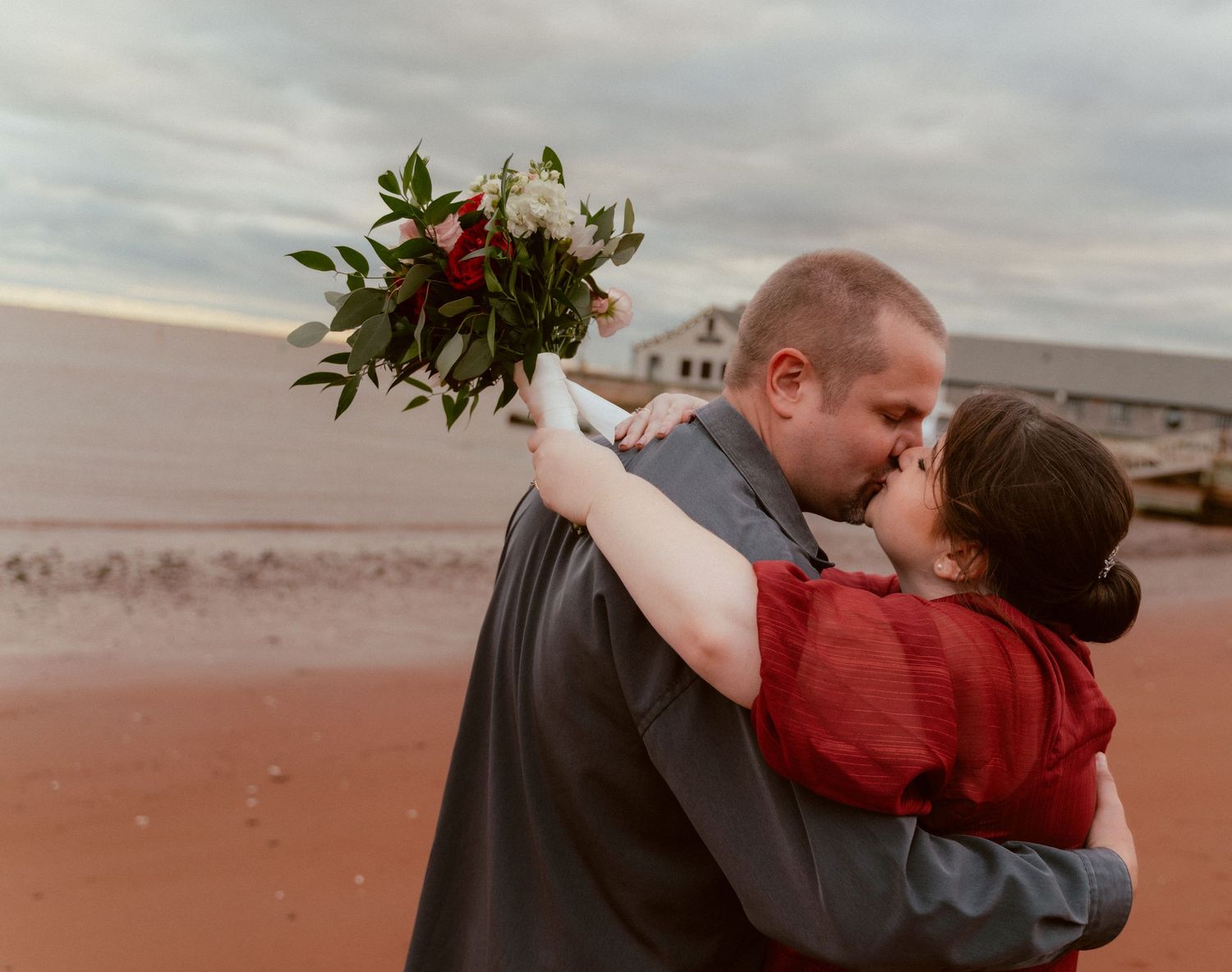 A couple shares a romantic kiss on a beach while holding a floral bouquet.