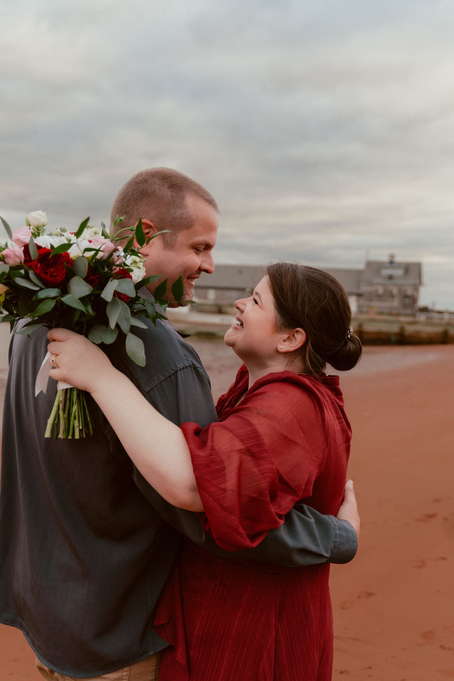 A romantic couple shares a joyful moment on a cloudy day, with one person holding a bouquet of red flowers.
