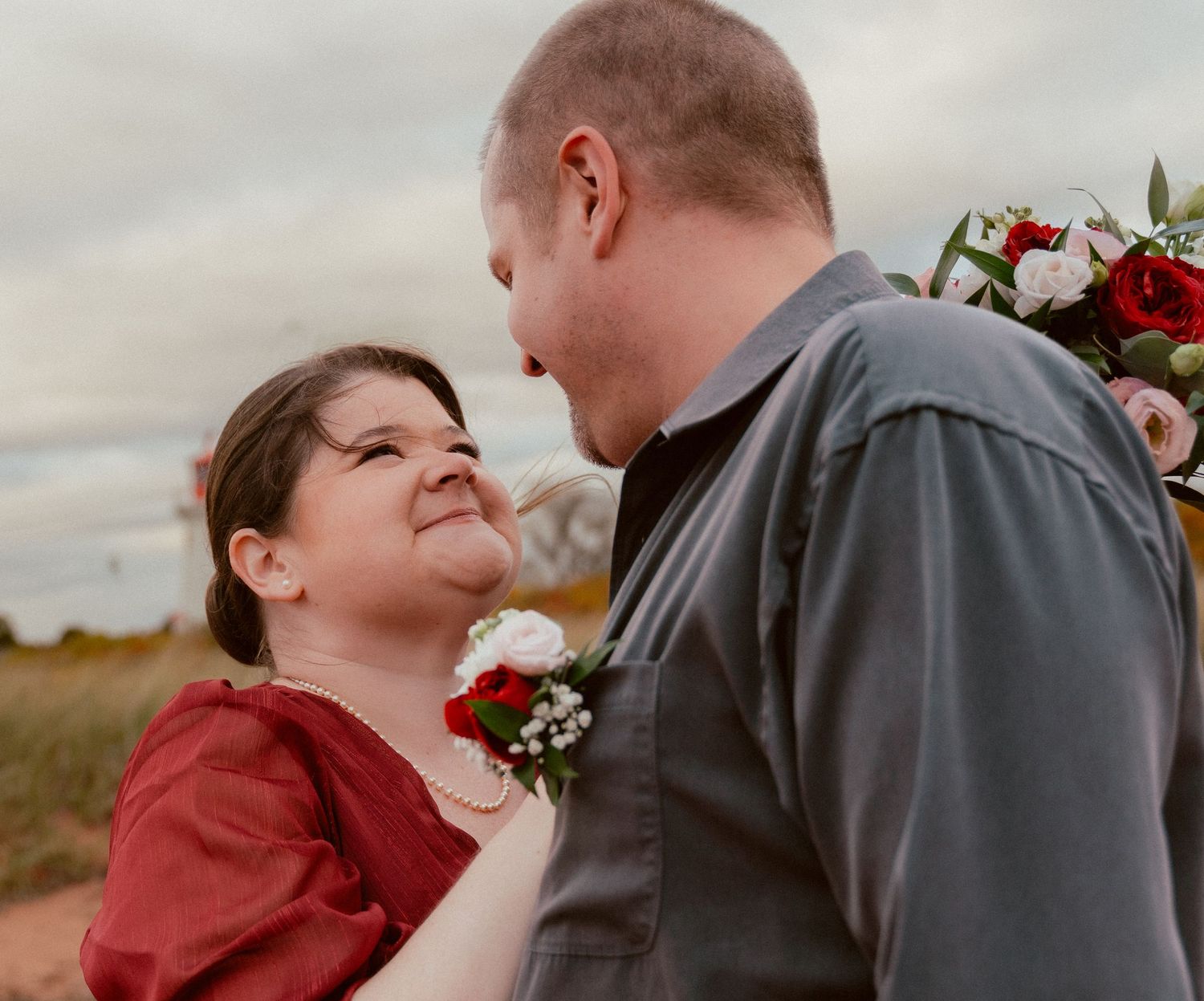 A close-up candid moment between a couple wearing red and grey, with flowers in hand against an overcast sky.