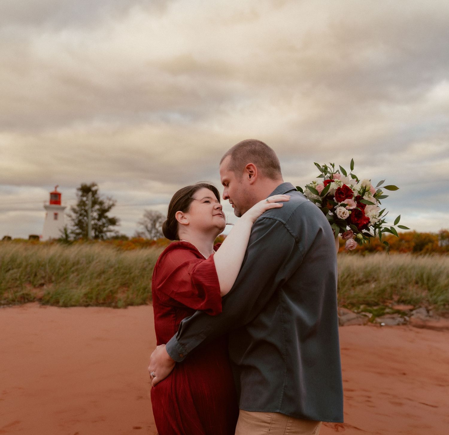 A romantic embrace near a lighthouse with red dress and floral bouquet details.