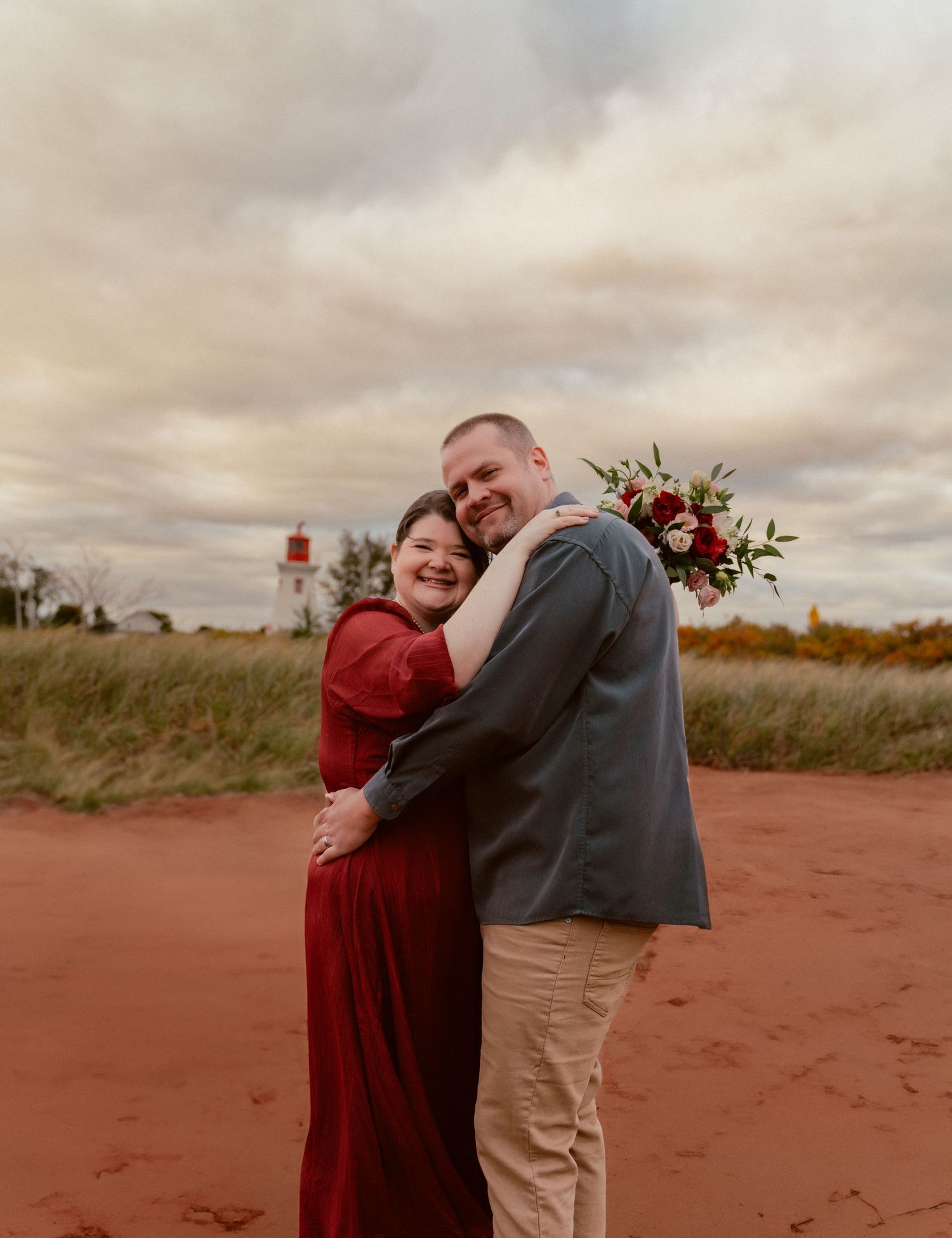 Couple embracing outdoors with lighthouse and autumn landscape in background.