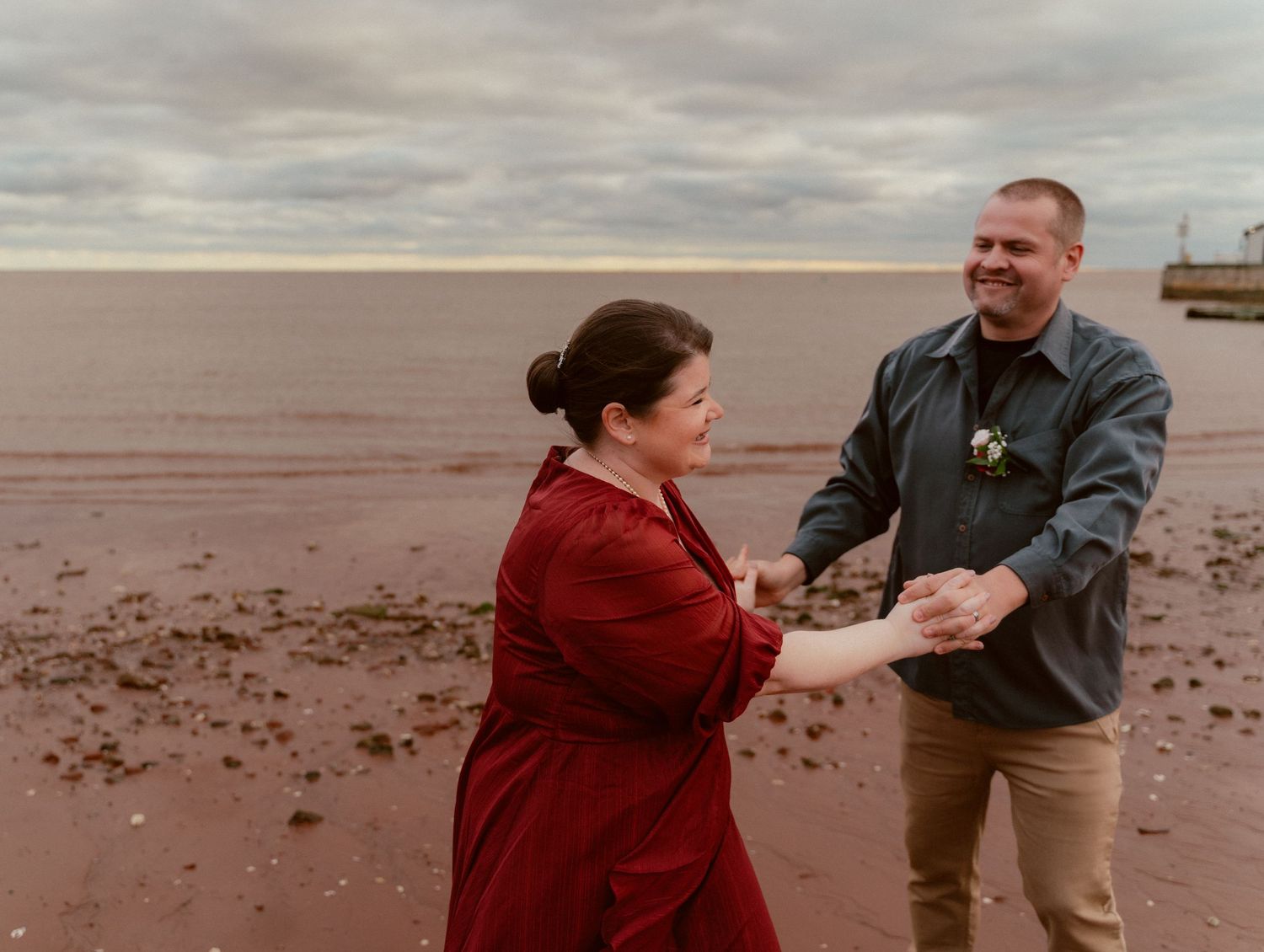 Two people dance together on a scenic beach under cloudy skies, with one wearing a red dress.