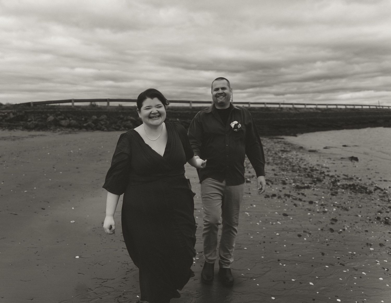 A black and white photo of a couple walking together on a beach under cloudy skies.