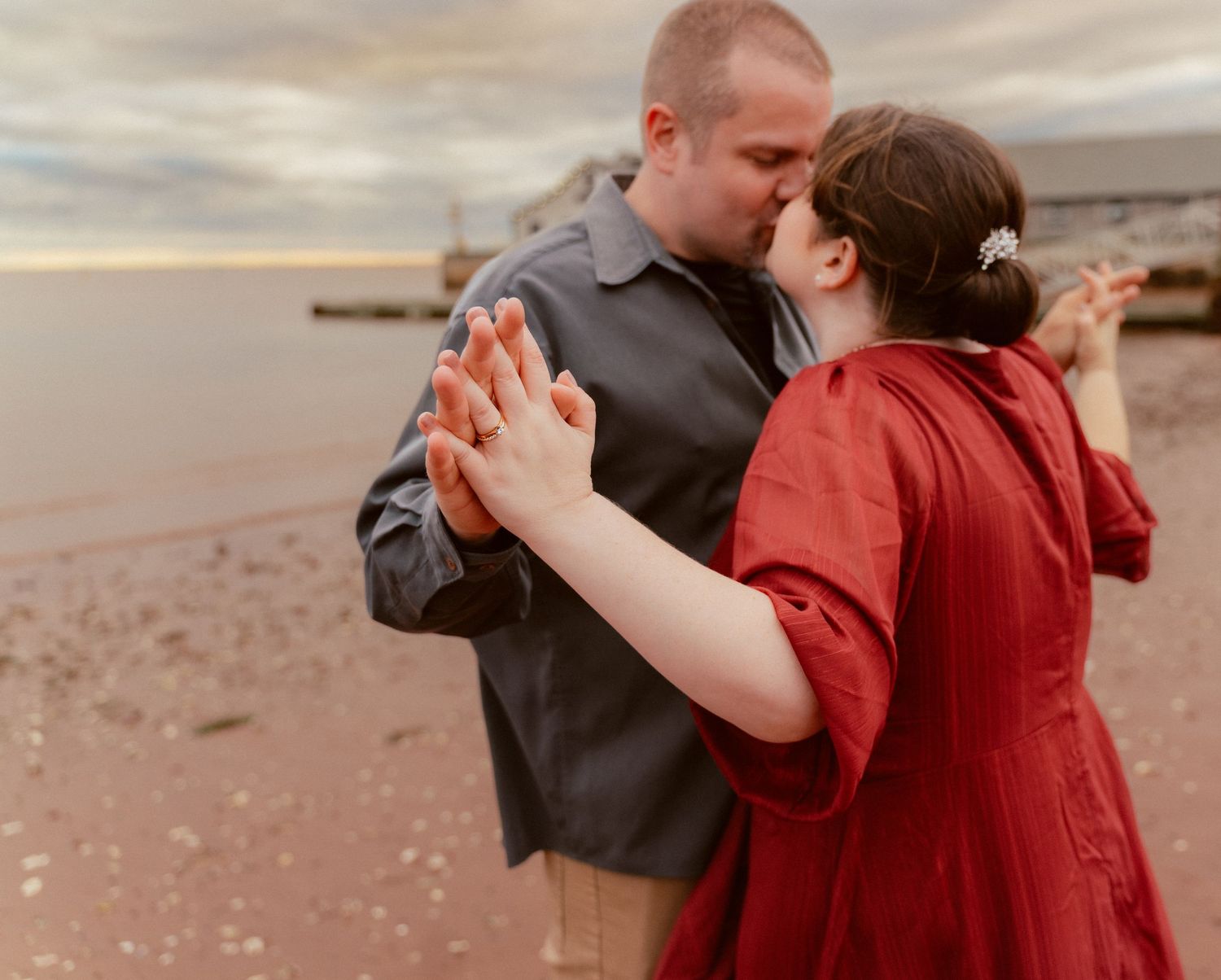 A couple dances on the beach at sunset, with one wearing grey and the other in a red dress.
