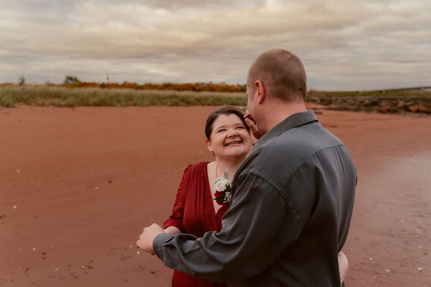 A couple embraces by a waterfront with cloudy skies, the woman wearing a deep red dress.