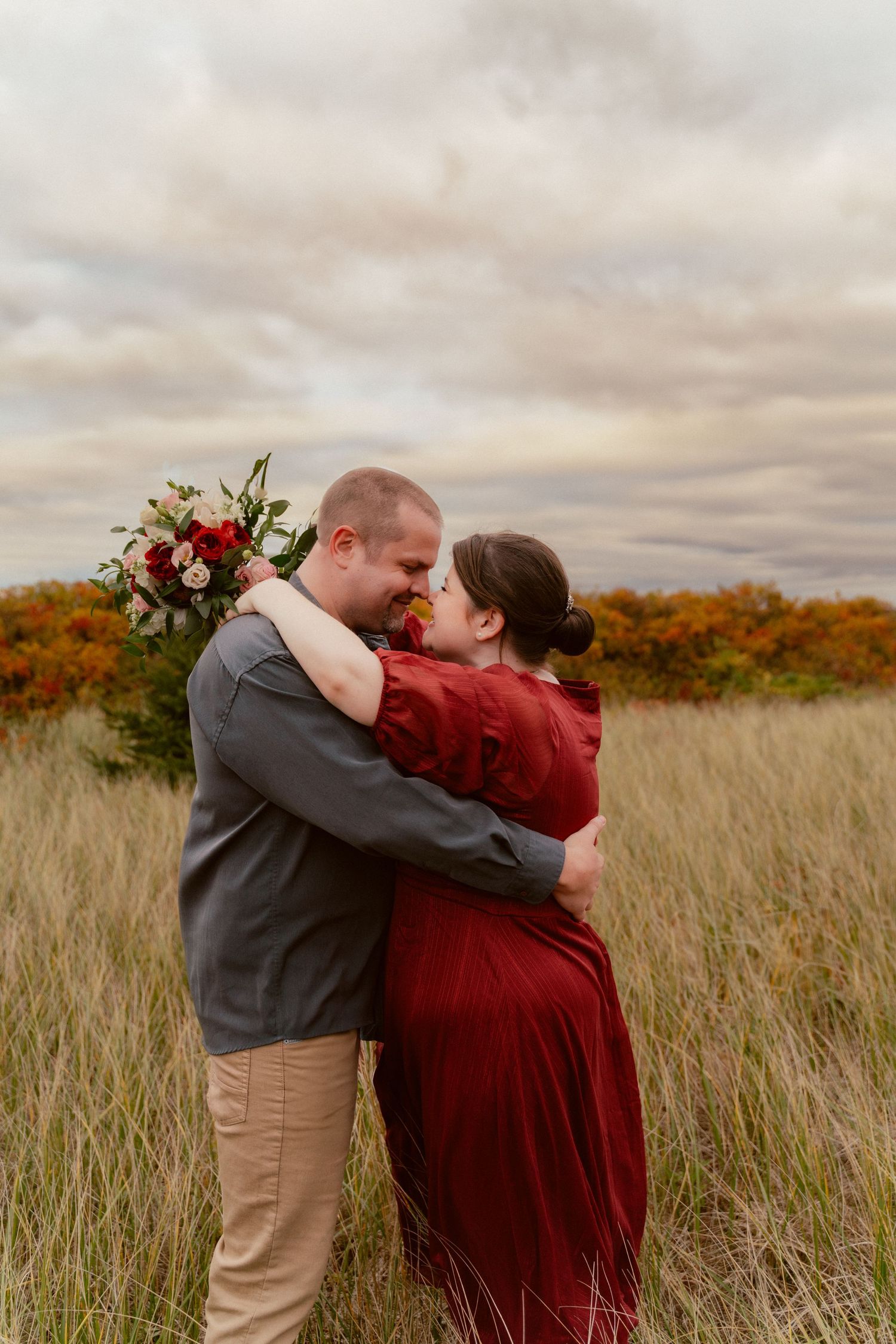 A romantic embrace in tall grass at sunset with autumn colors and a floral bouquet.