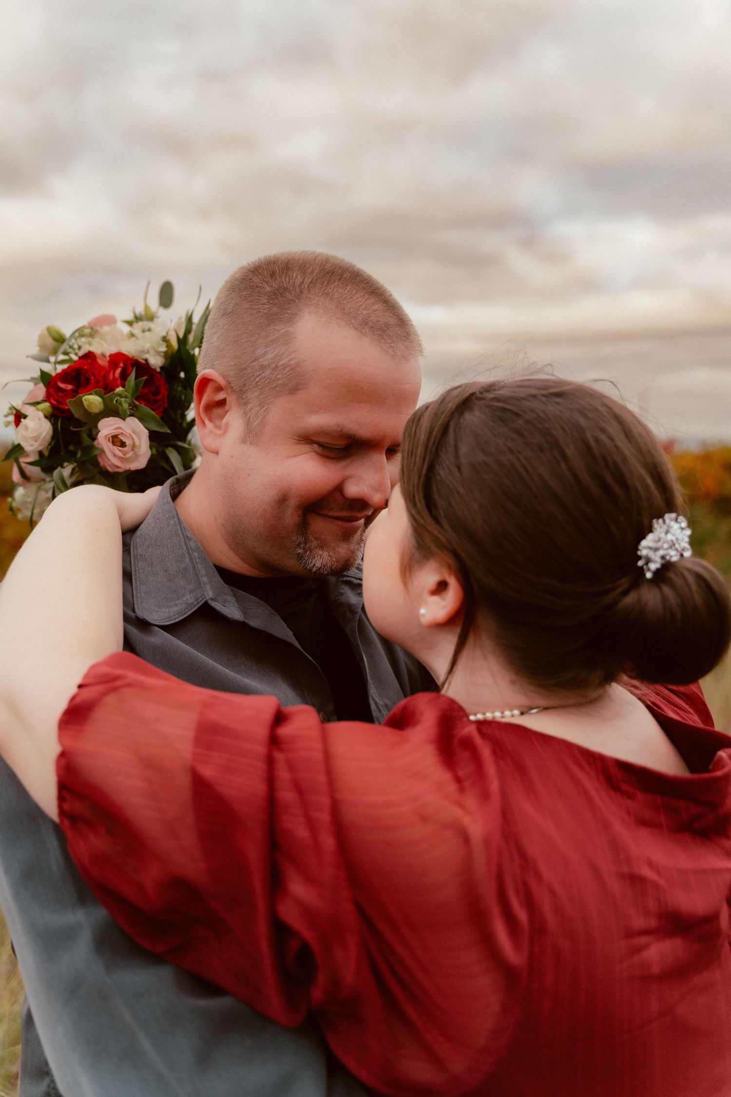 Couple embraces outdoors with red and pink flower bouquet against cloudy autumn sky.