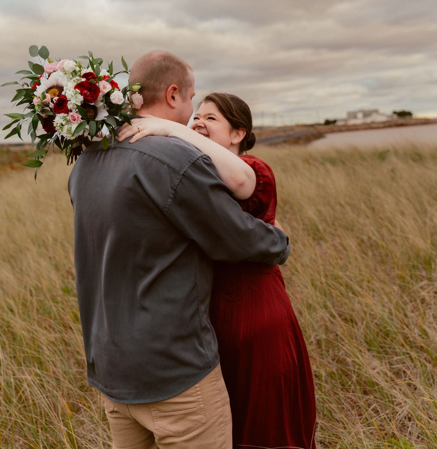 A romantic moment captured in tall grass with the woman holding a red and white floral bouquet.